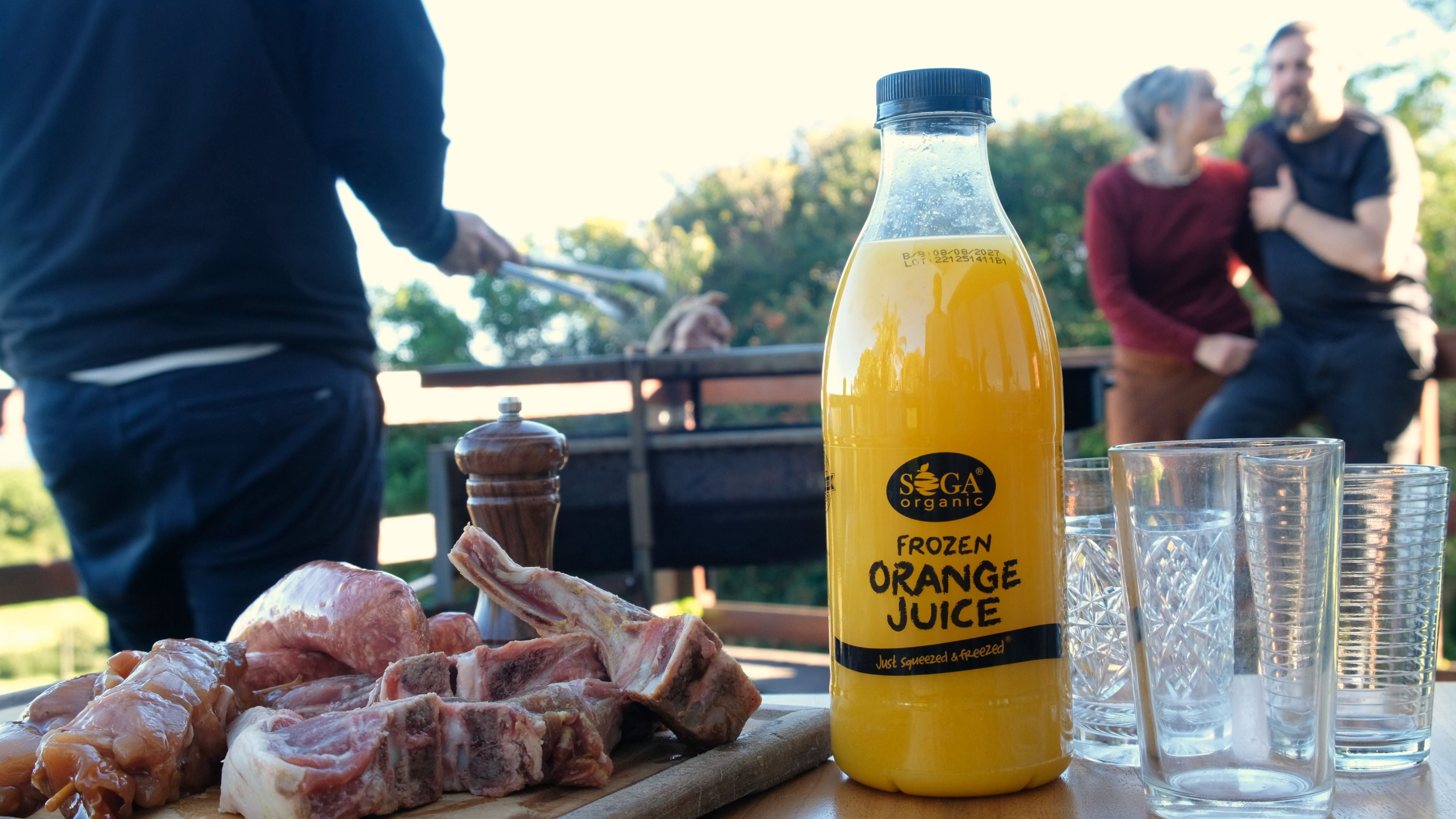 SOGA frozen orange juice bottle on a deck table beside raw meat, with a couple and a braai behind
