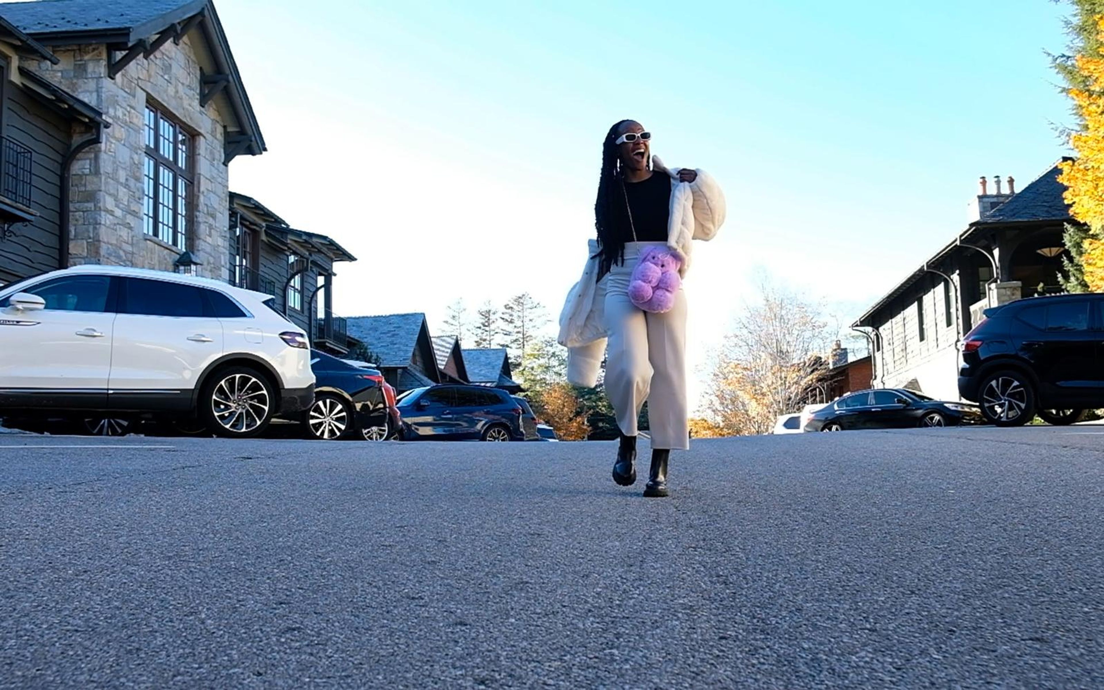 Wide low shot of model approaching along an empty road, white jacket and cream trousers, houses and cars
