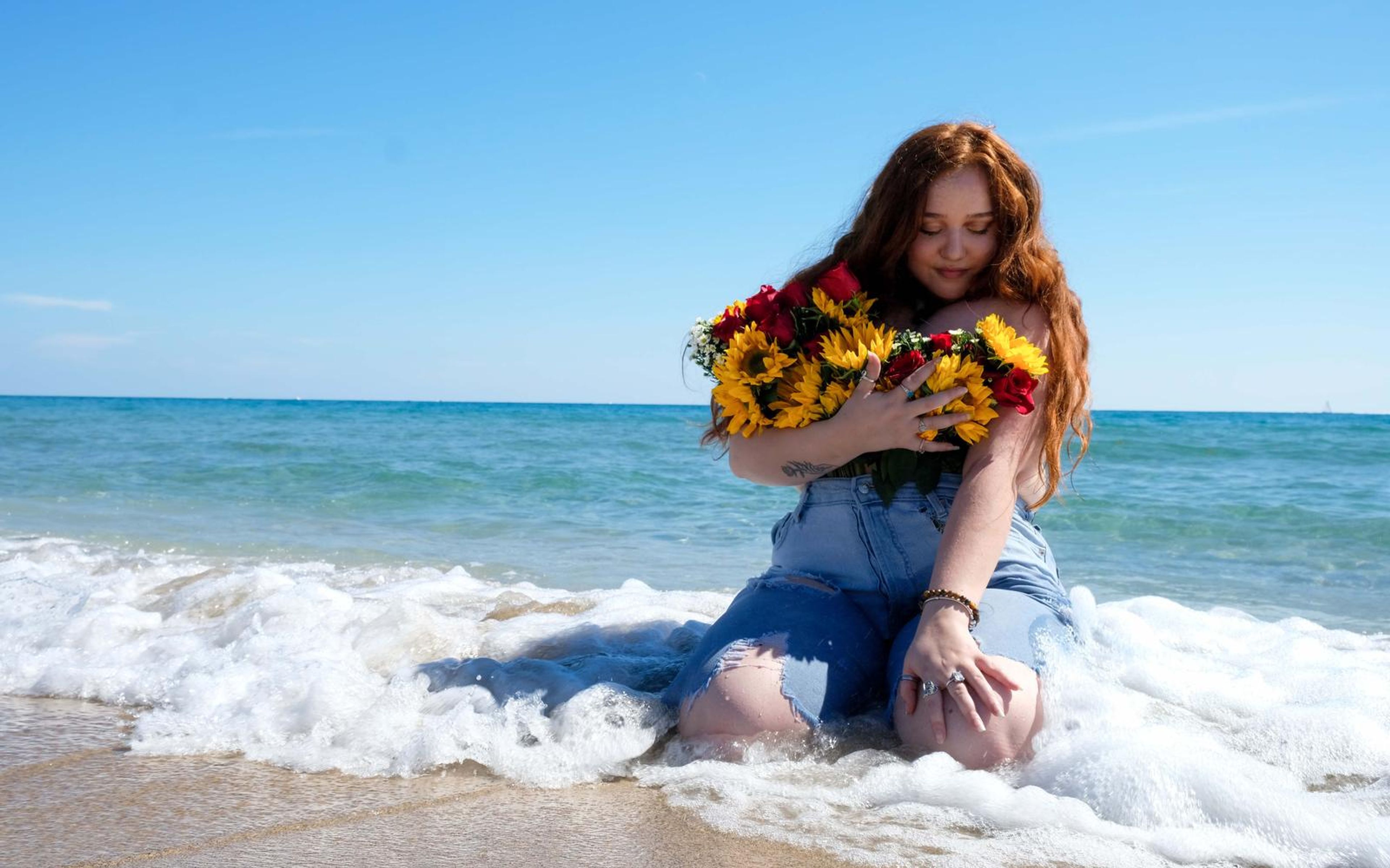 Model seated in bright surf with the bouquet in her arms, wave foam spreading around her denim