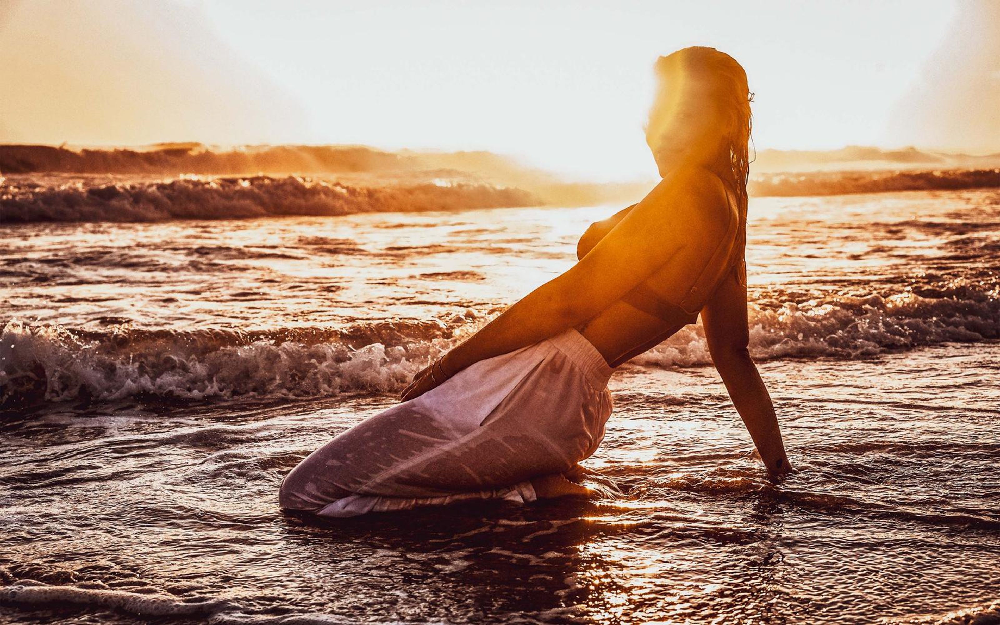 Woman leaning back on one arm while kneeling in the surf, sun flare across her face, warm orange sky