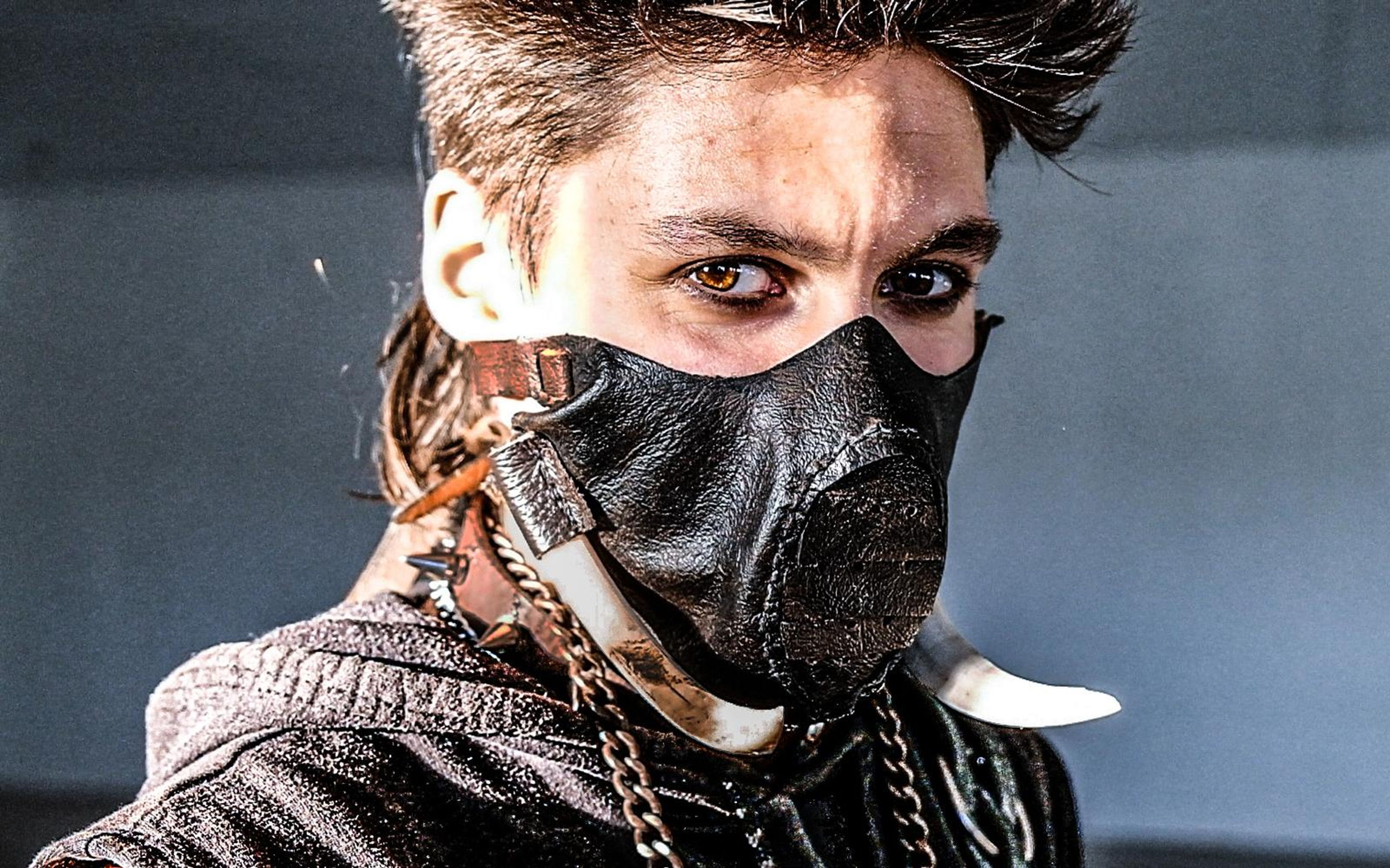 Close portrait of a masked figure with spiky hair in a leather face mask with a curved tusk and chain, grey backdrop