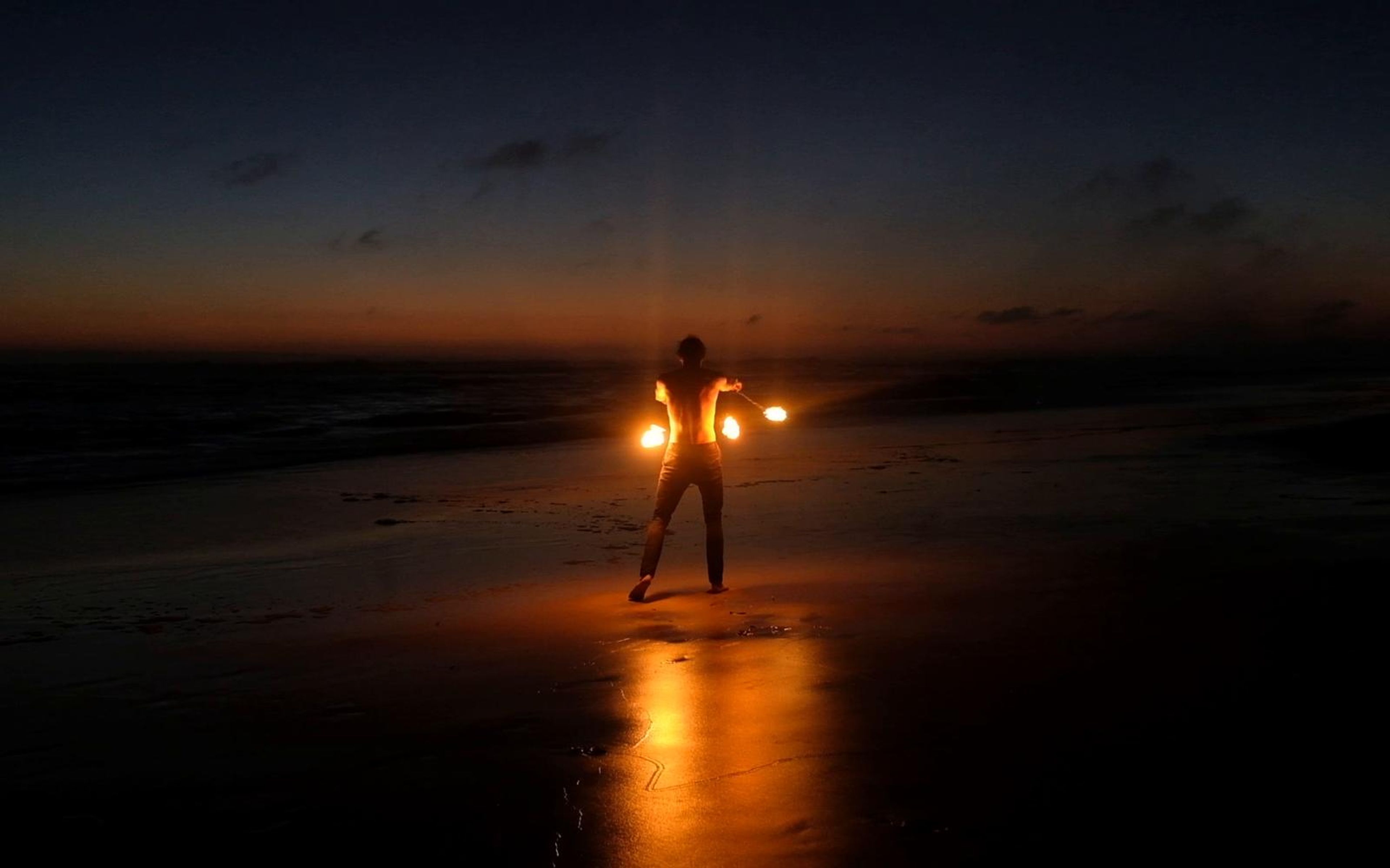 Performer silhouetted on the beach at dusk with flames reflecting on wet sand
