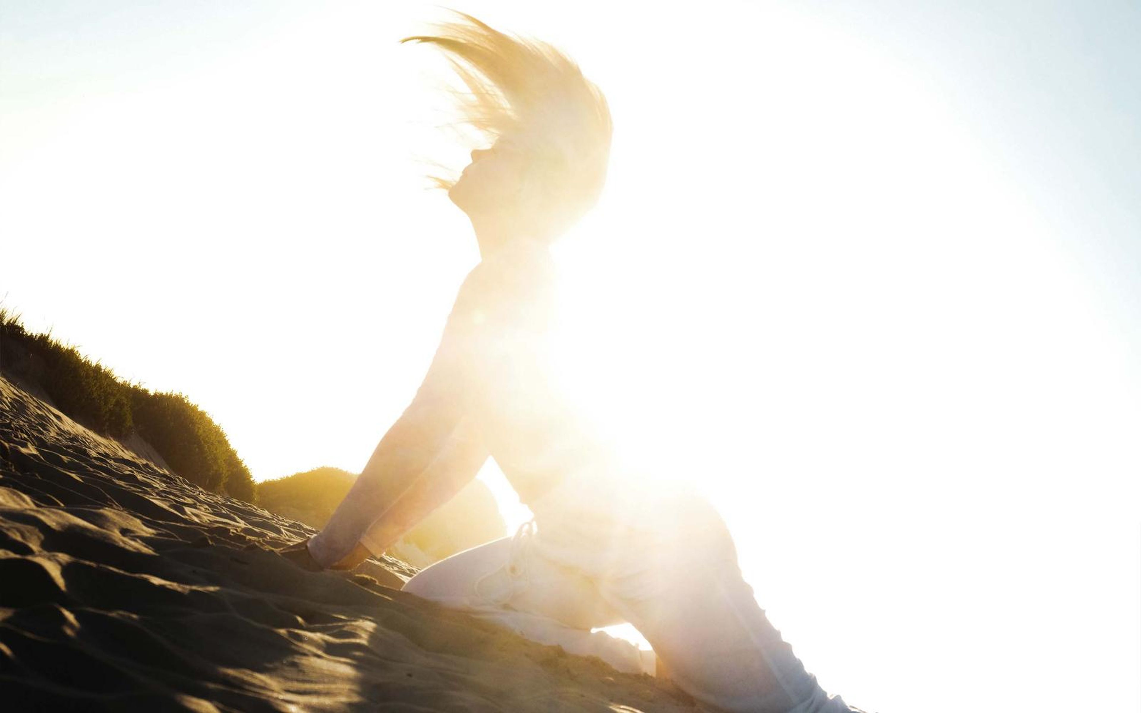 Woman kneeling on a dune with hair thrown back, almost lost in blown-out sunlight