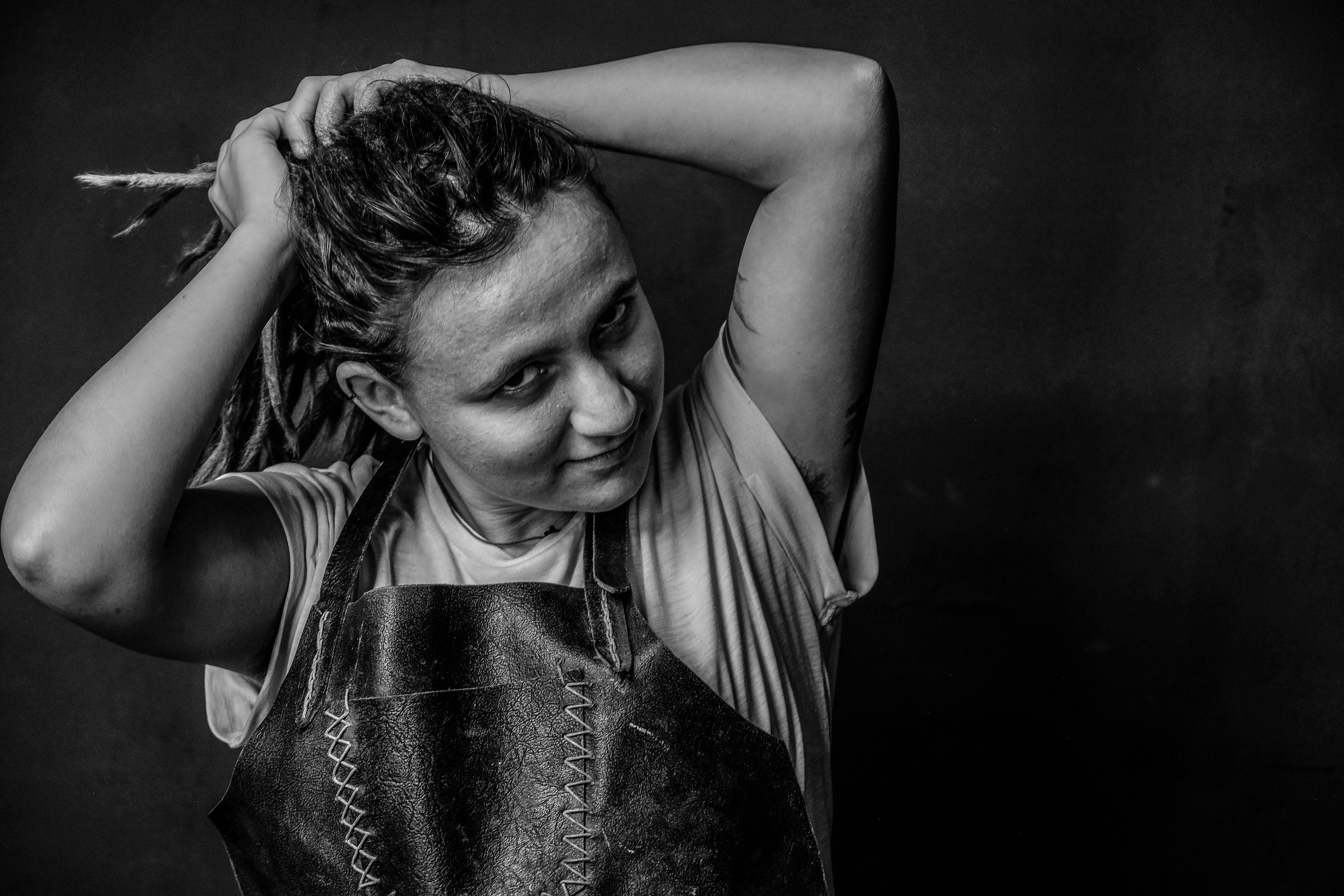 Black and white portrait, subject gathering dreadlocks up with both hands, wearing a leather apron