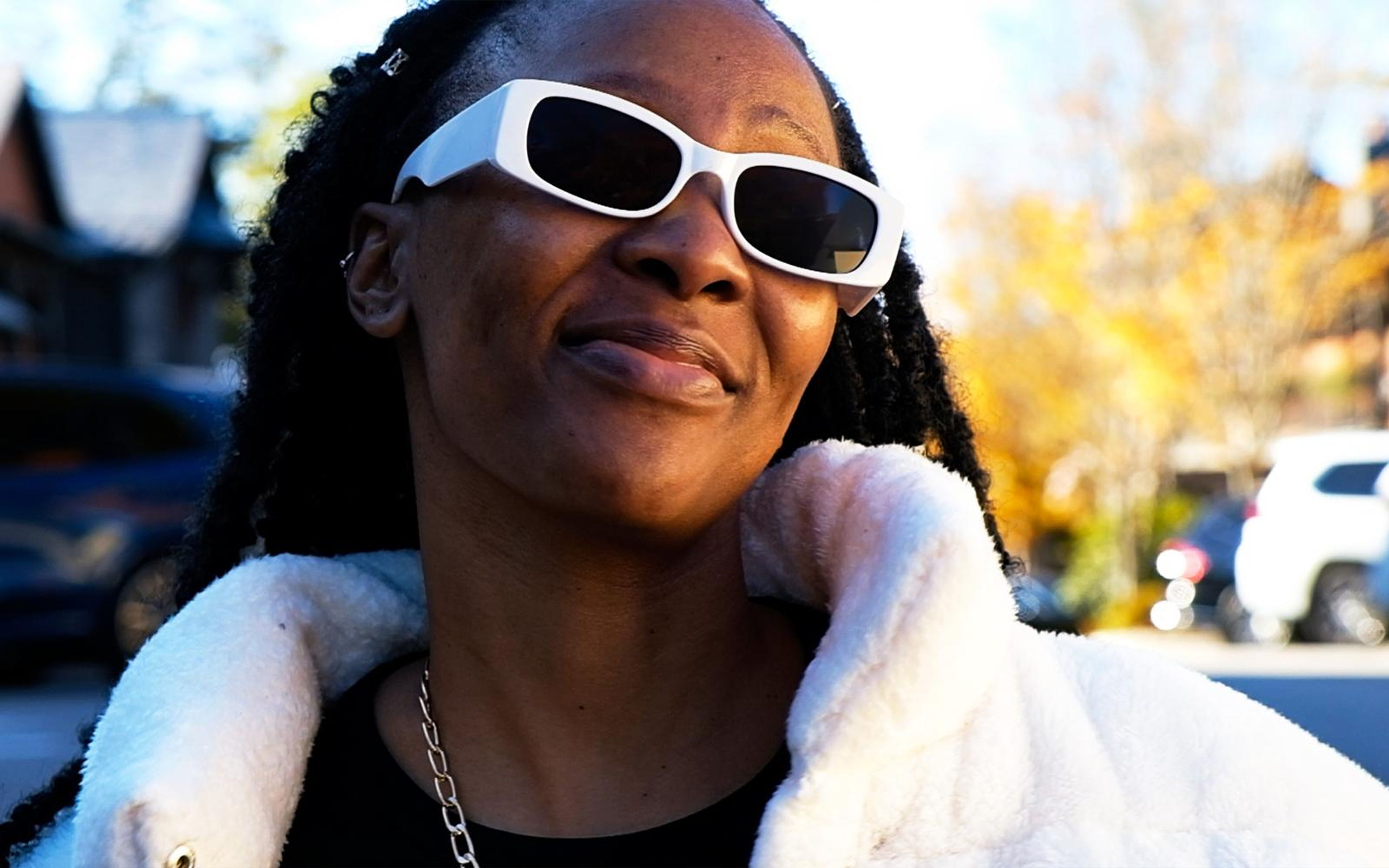Model in white sunglasses smiling and tilting her head, fingers at her fur collar, village street