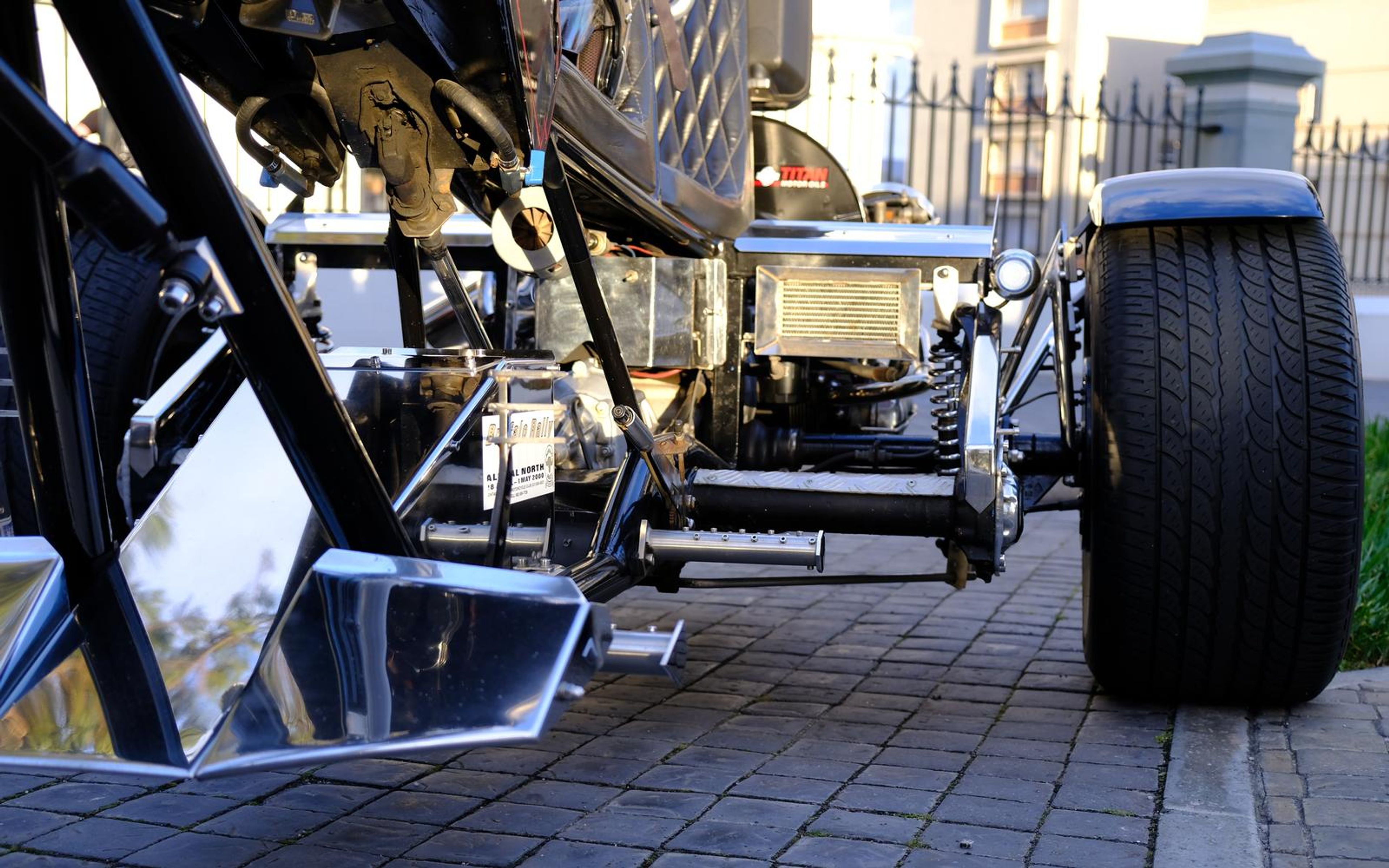 Underside view of a trike frame with chrome parts and a wide tyre, buildings behind