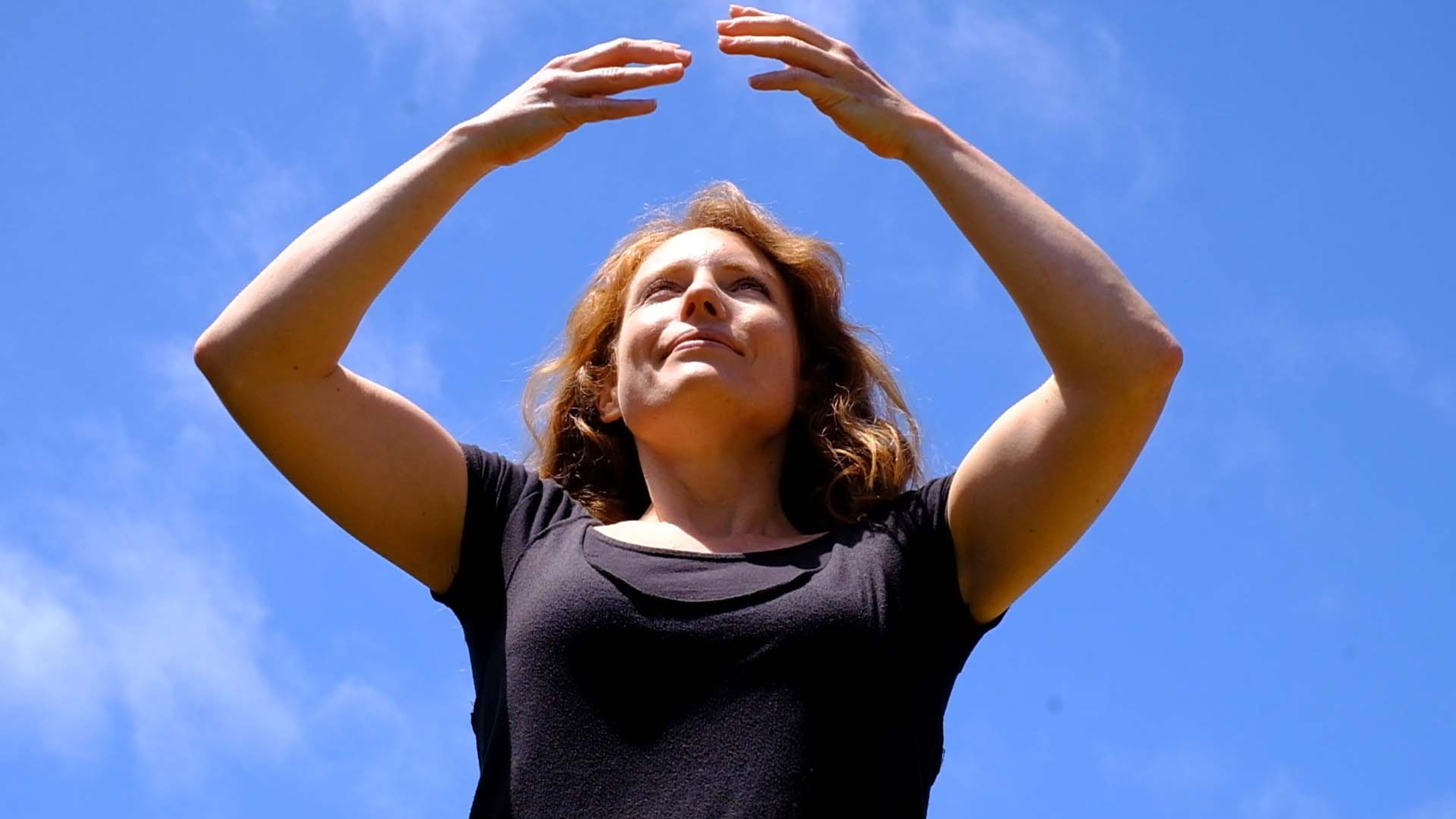 Woman with arms raised overhead against a bright blue sky during an outdoor movement session