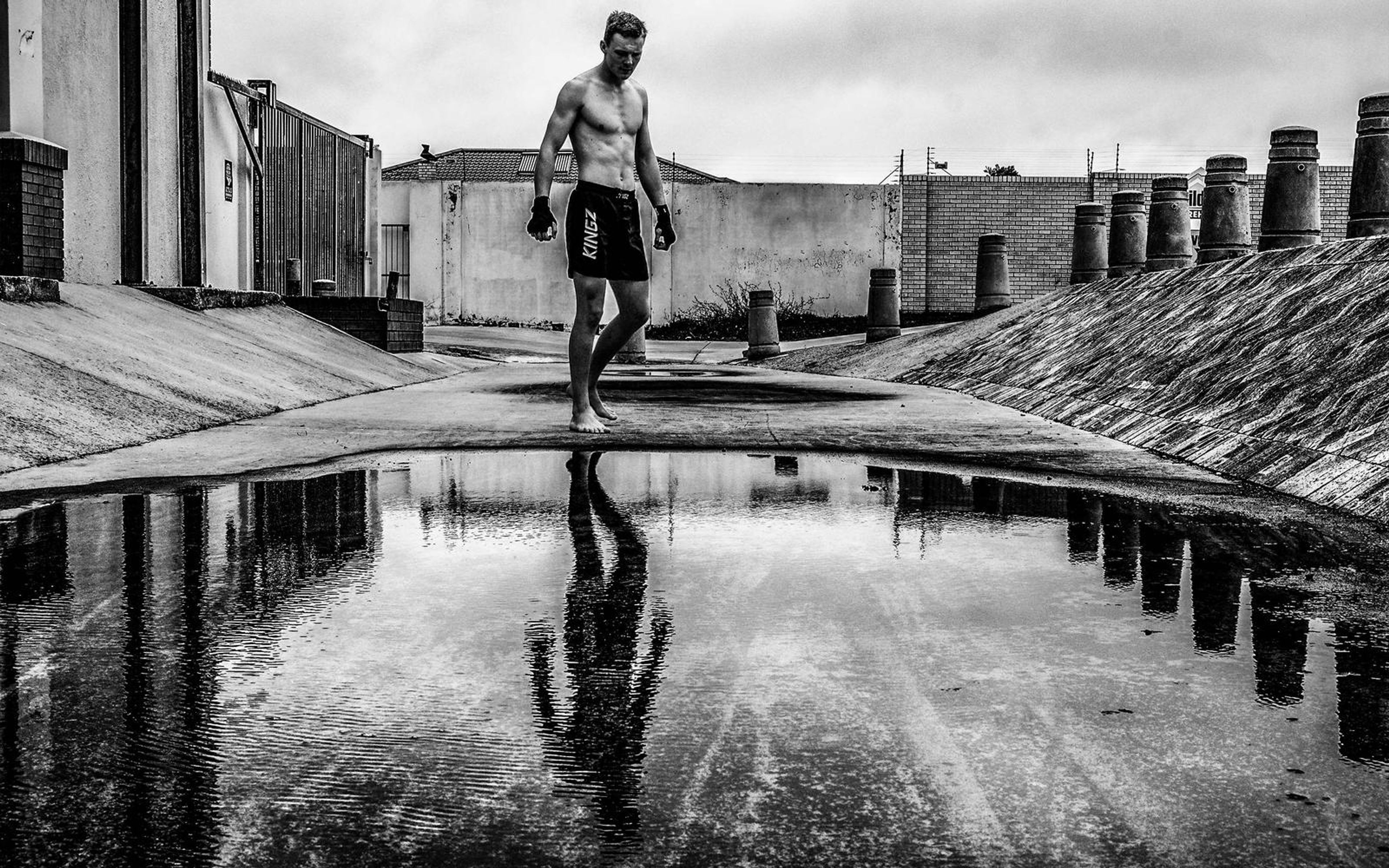 Black and white image of a man standing by a reflective puddle, concrete embankment behind him