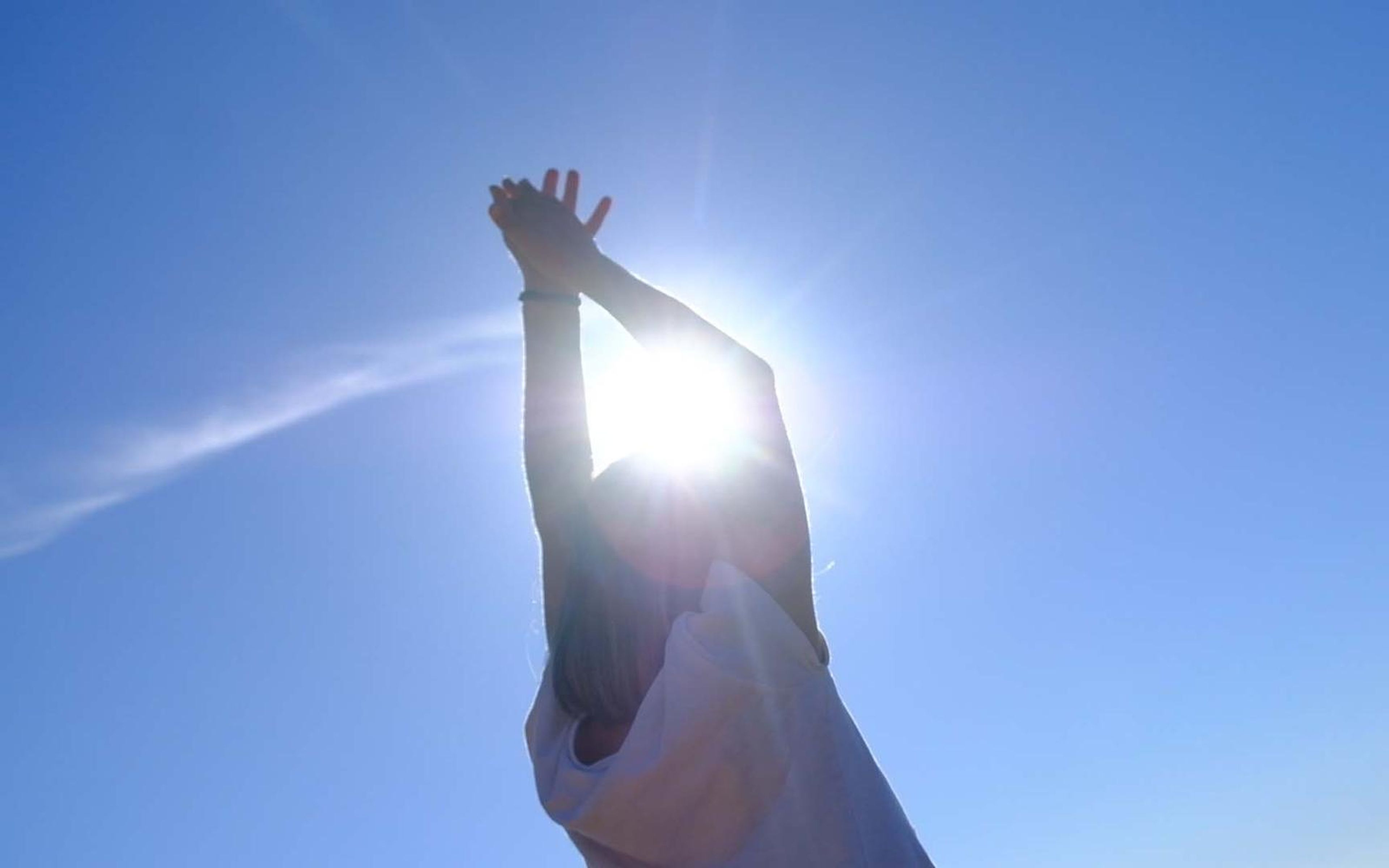 Raised hands against a clear blue sky with the sun flaring between the fingers