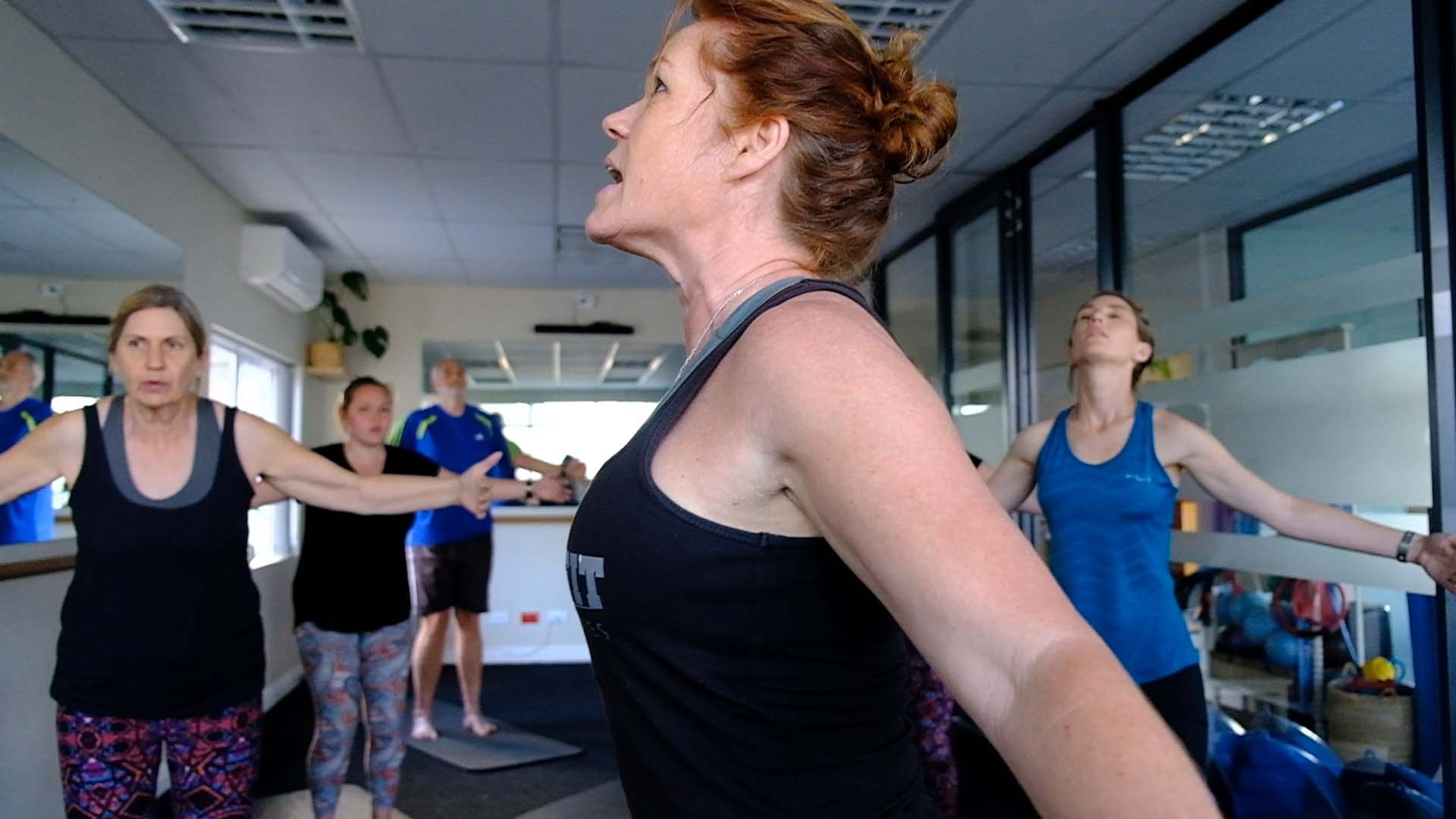 Women in a group class holding a side stretch, one in a black tank top looking up