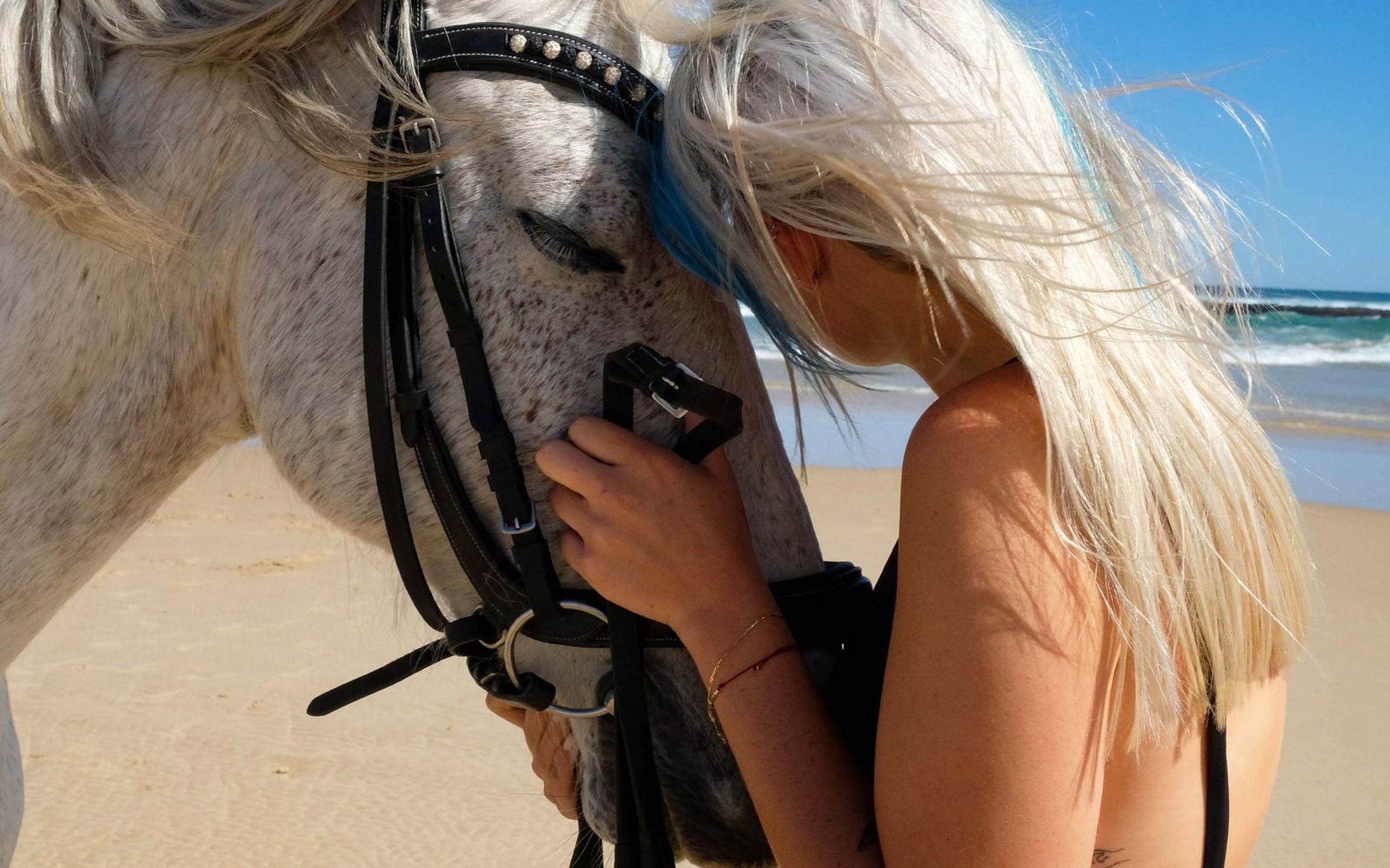 Woman in a black swimsuit holding a horse's bridle while hair blows across her face