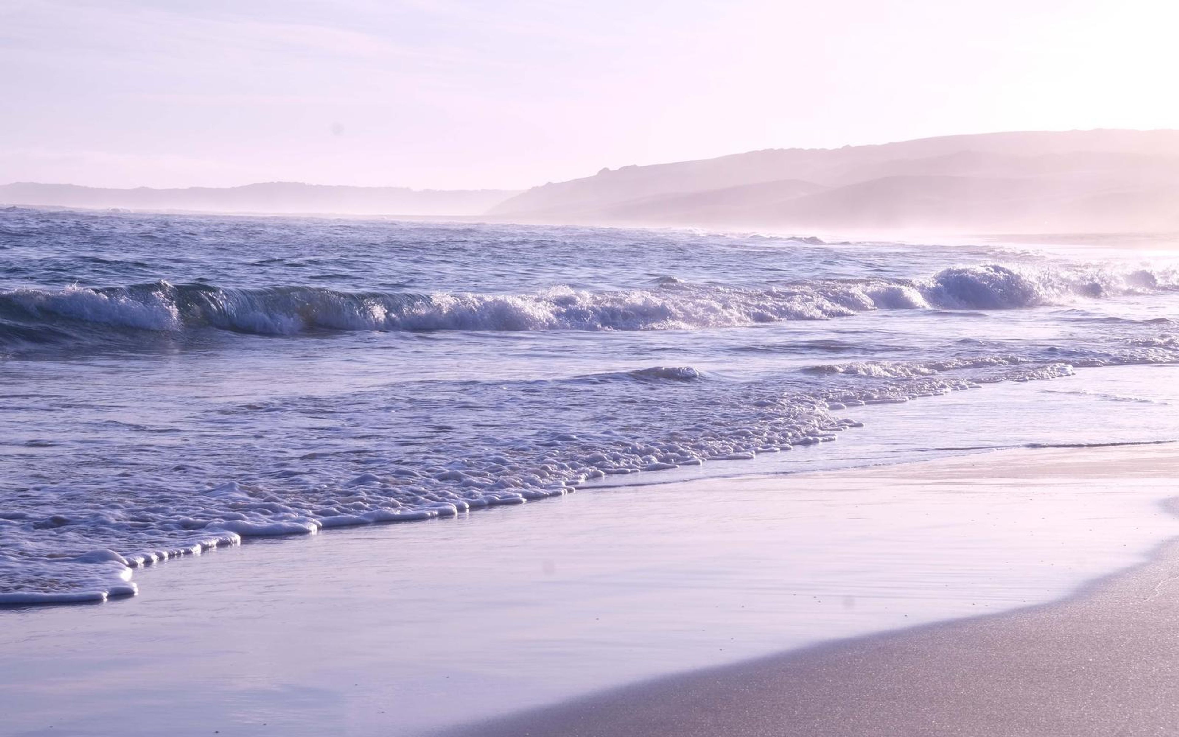 Waves washing onto a pale beach in soft light with misty headland in the distance