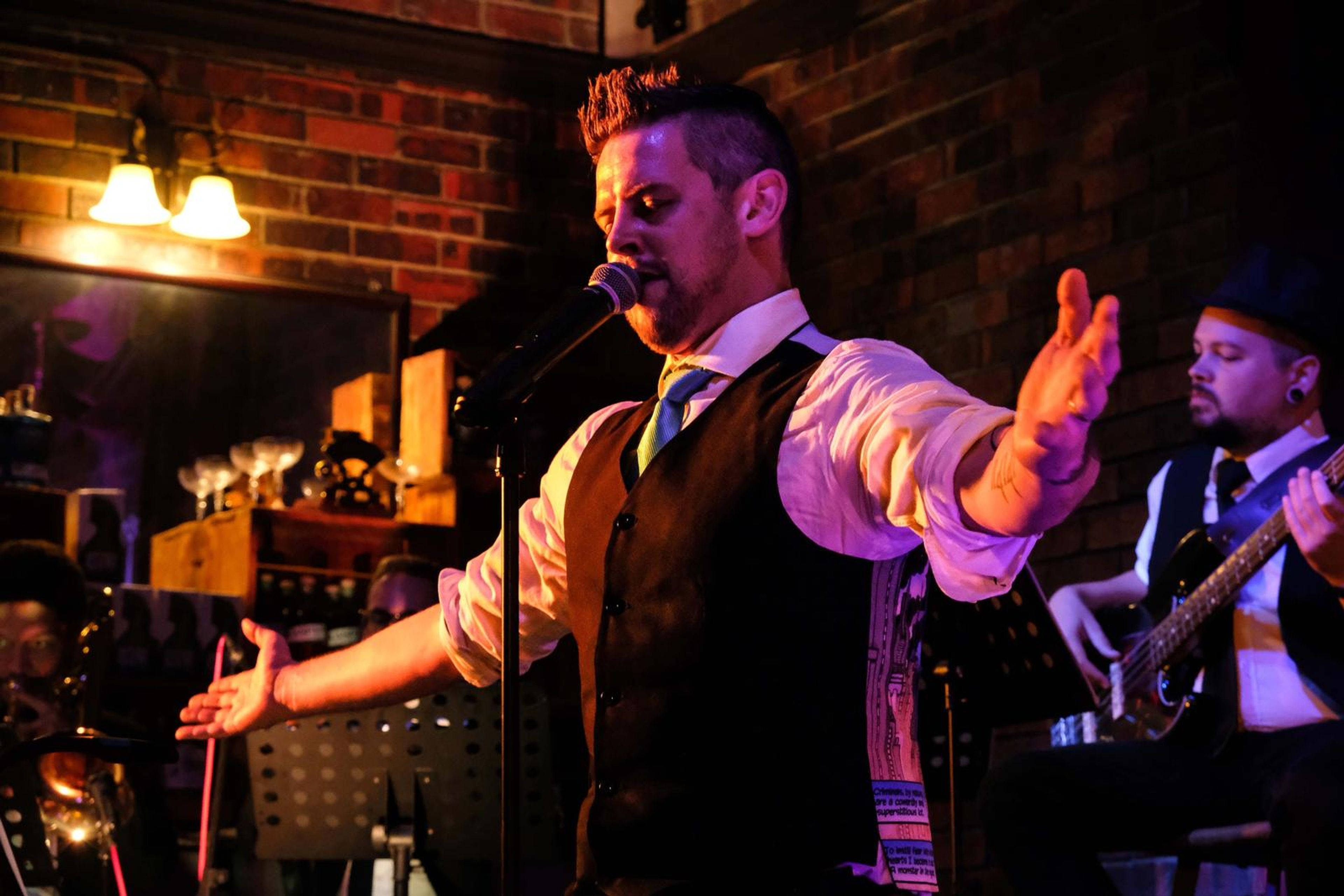 Lead singer in a waistcoat with his arms spread wide in front of a brick wall at Nineteen 33