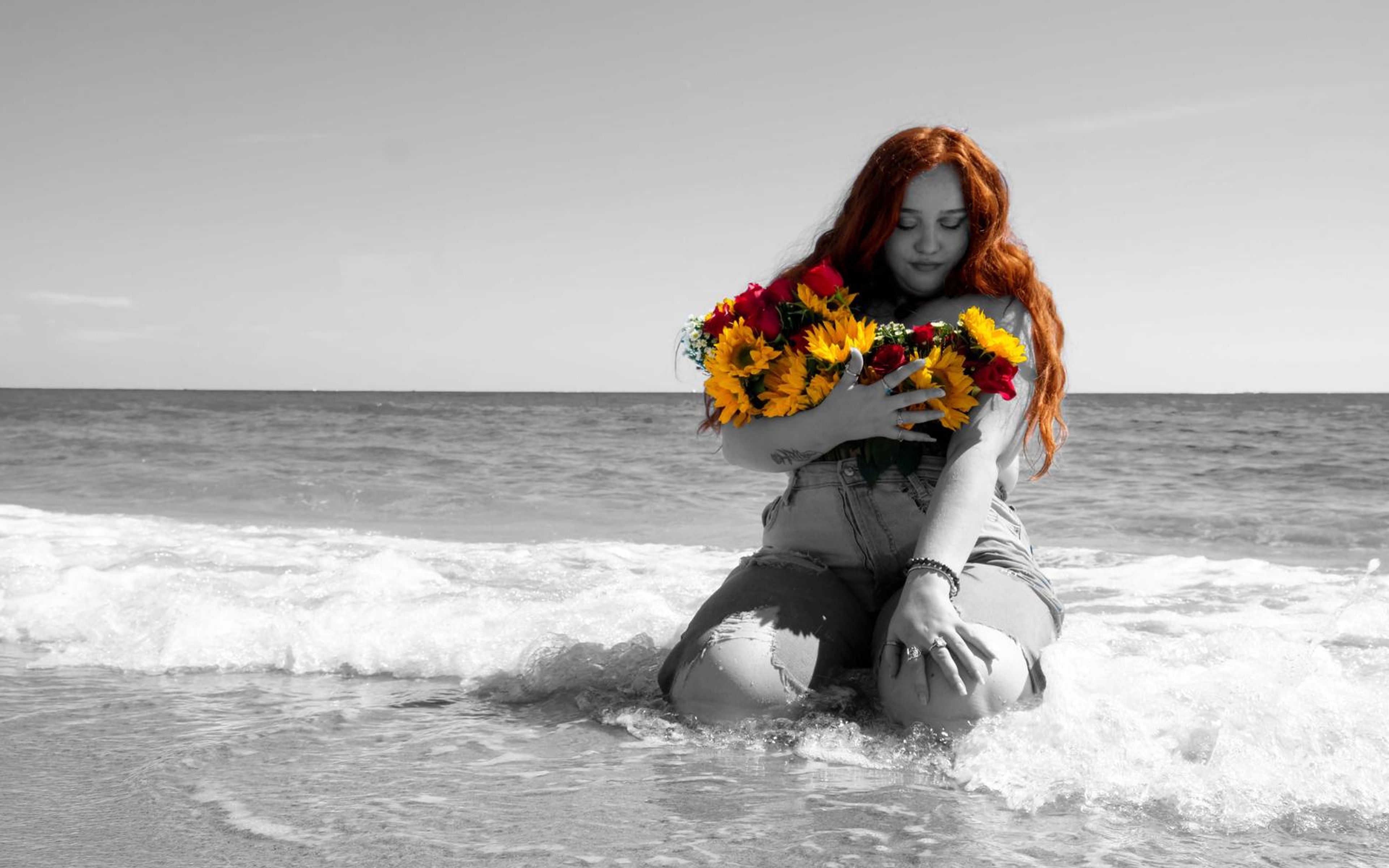 Black and white beach portrait, model kneeling in rushing foam, head tilted down, bouquet in colour