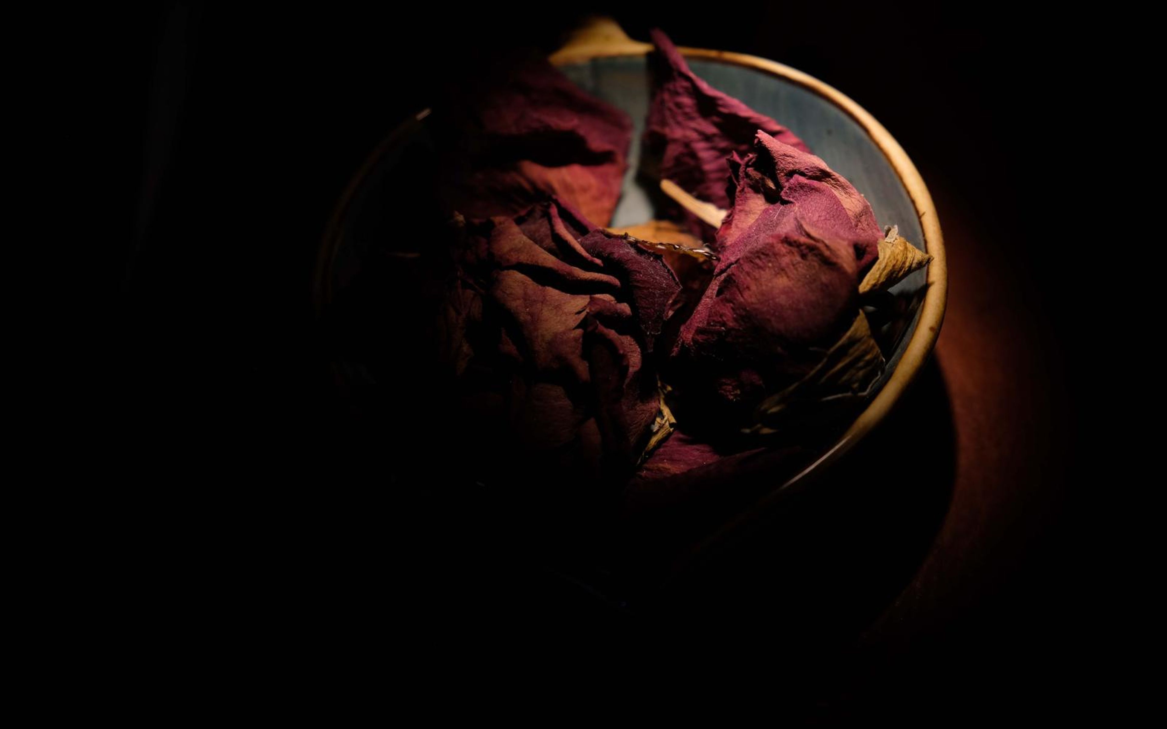 Dried red roses in a shallow bowl, lit with a warm glow against black
