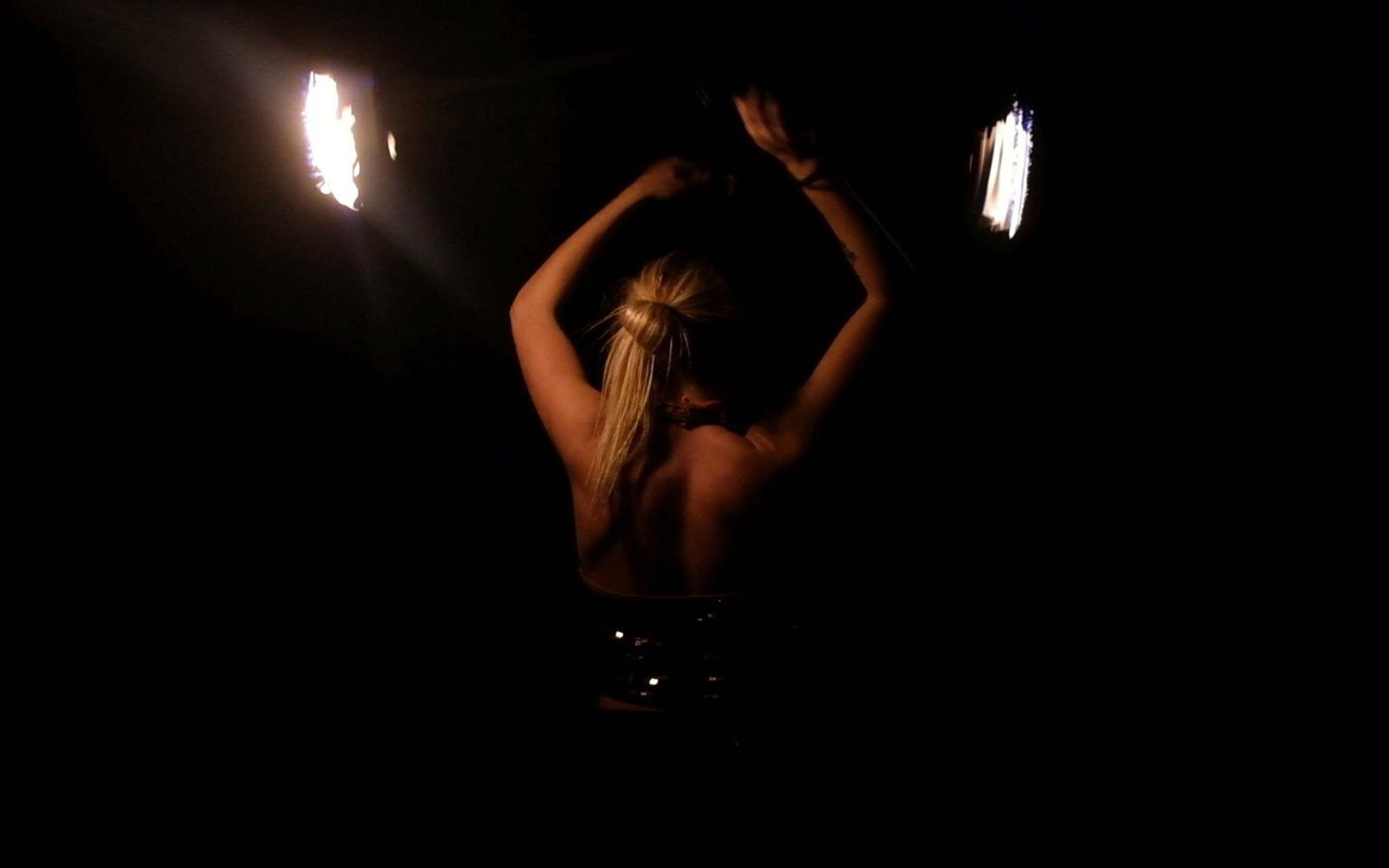 Performer seen from behind with arms raised overhead and fire streaks at both sides in a dark space