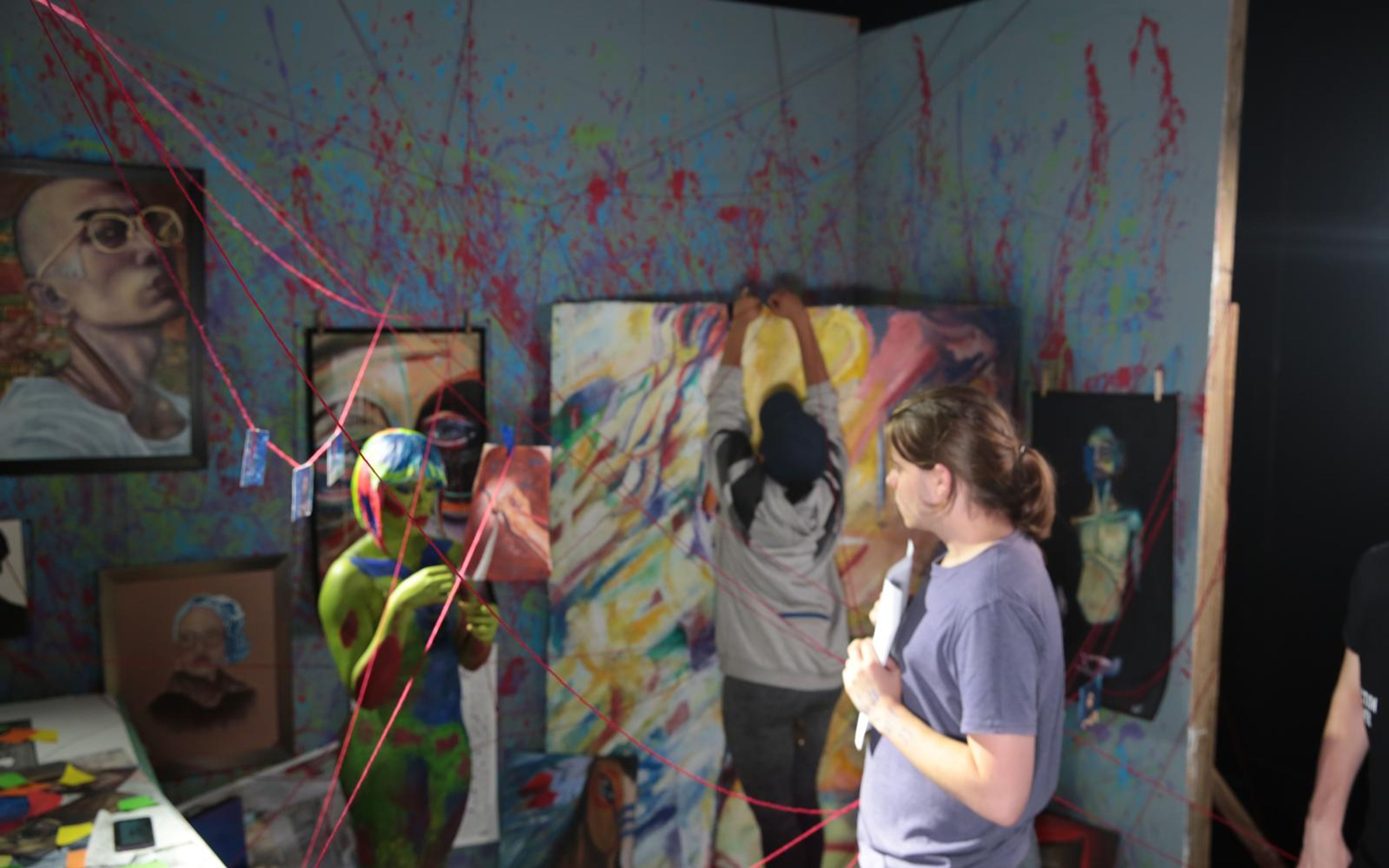 Person painting on a large canvas inside the art room set, with portraits and red strings across the walls