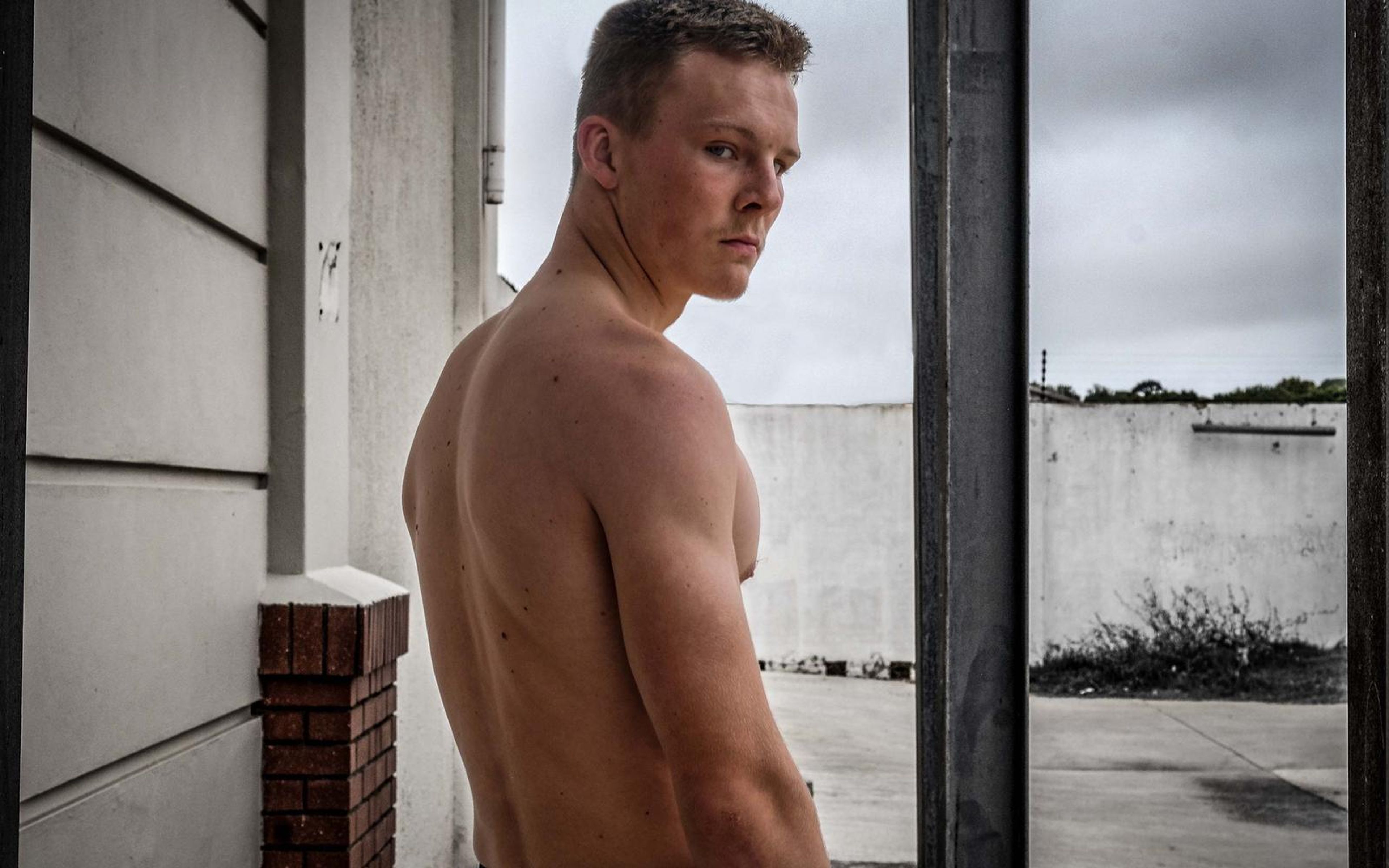 Warm-toned portrait of a man glancing over his shoulder, bare back to camera, grey sky through the opening