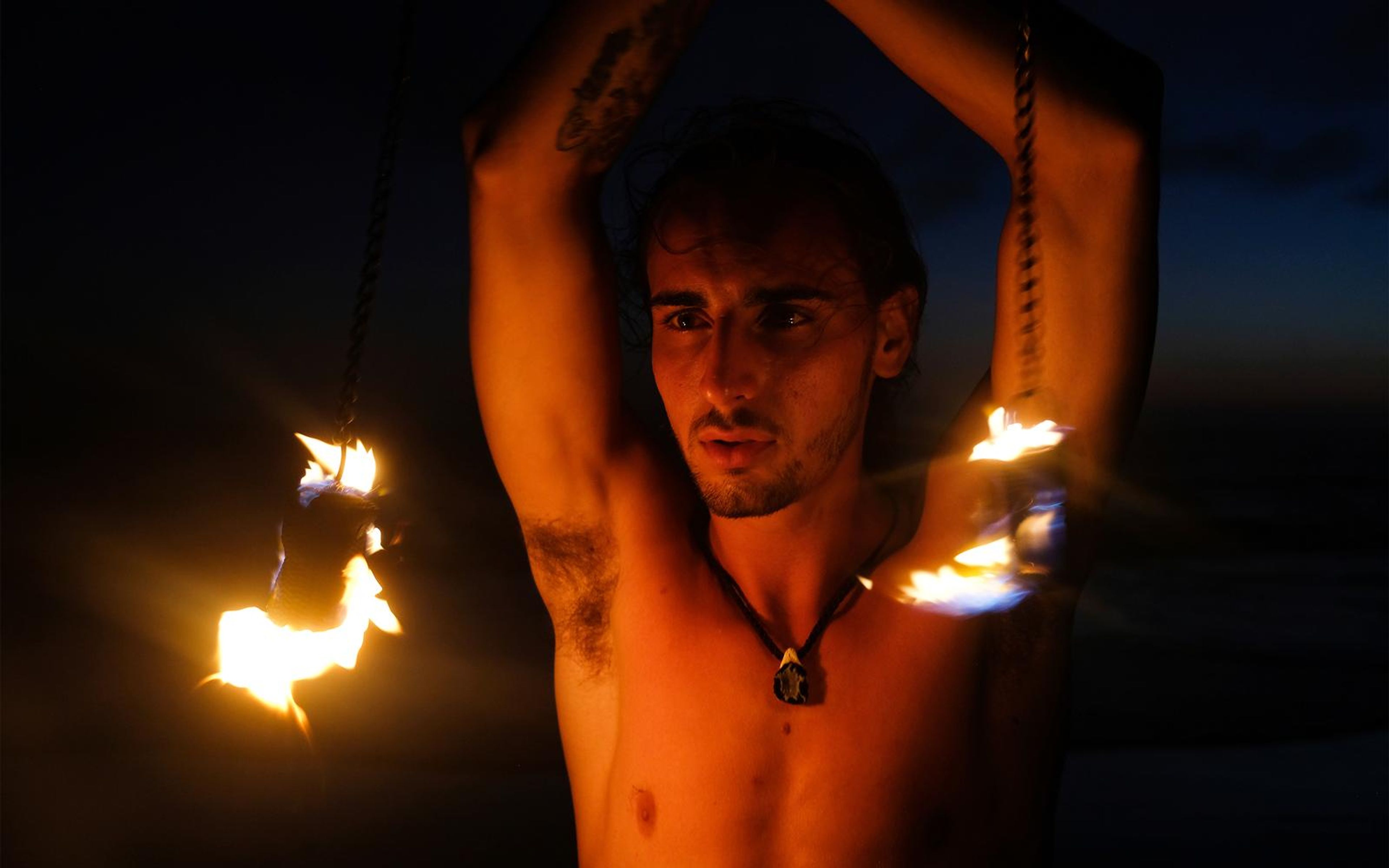 Man with both arms raised overhead and fire poi flaring at each side, dark blue dusk sky behind