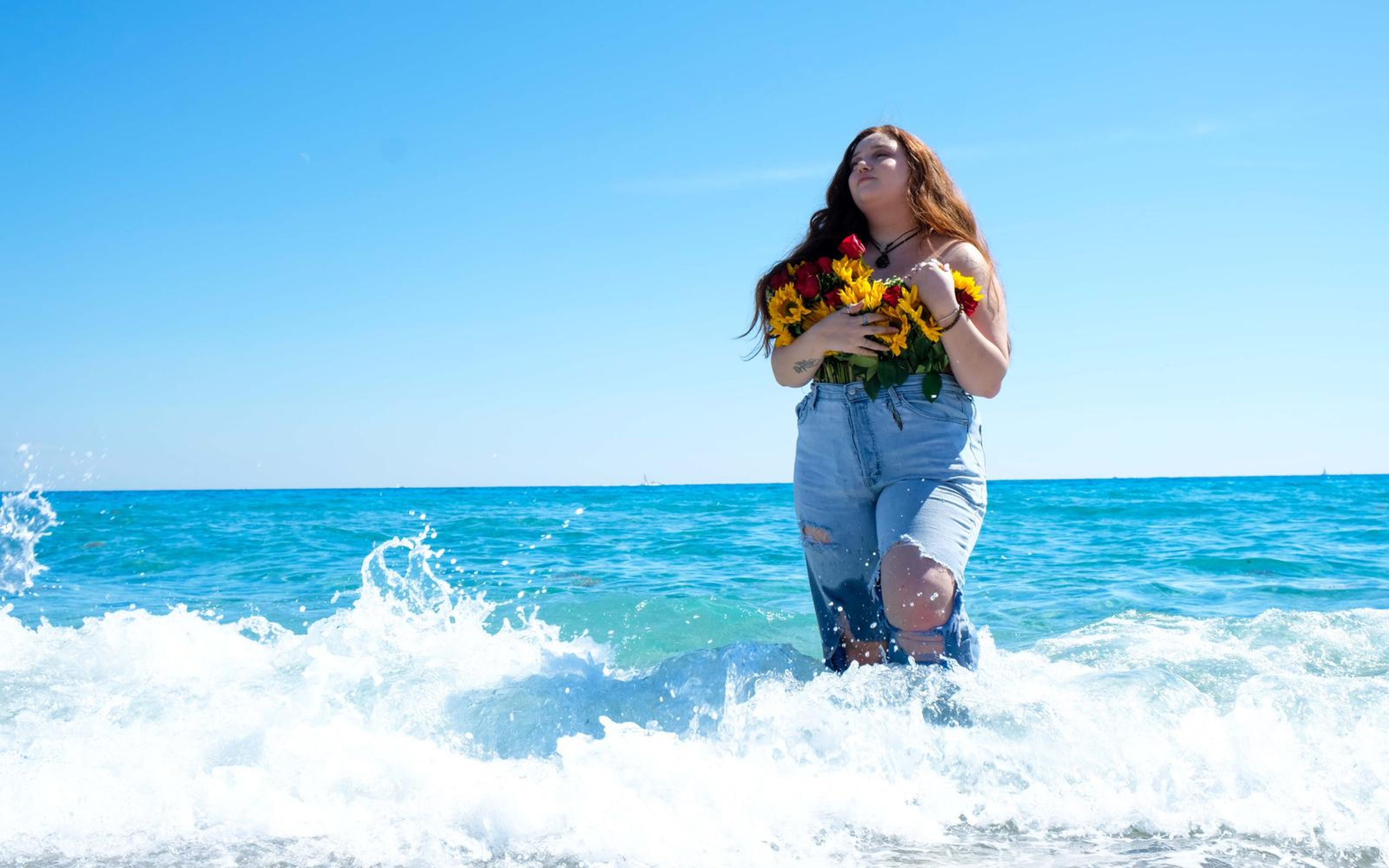 Model looking up in the surf as a wave crashes behind her, bouquet in arms, sunny blue sky