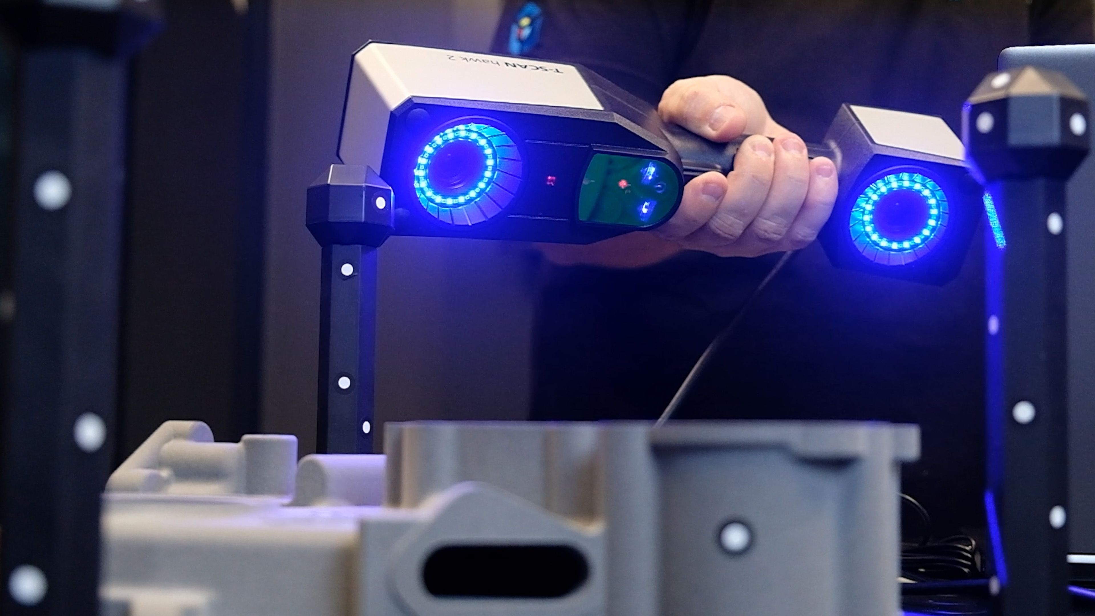 A hand holds the ZEISS T-SCAN Hawk 2 handheld 3D scanner, its blue LED rings lit, scanning a grey industrial part mounted on a photogrammetry rig