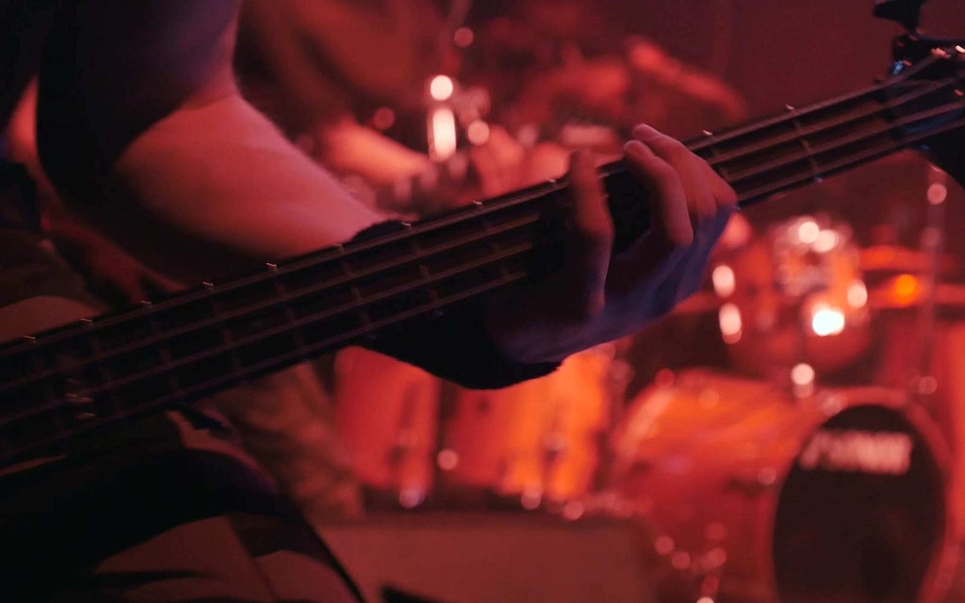 Bass player's fretting hand on the neck with a red-lit drum kit behind