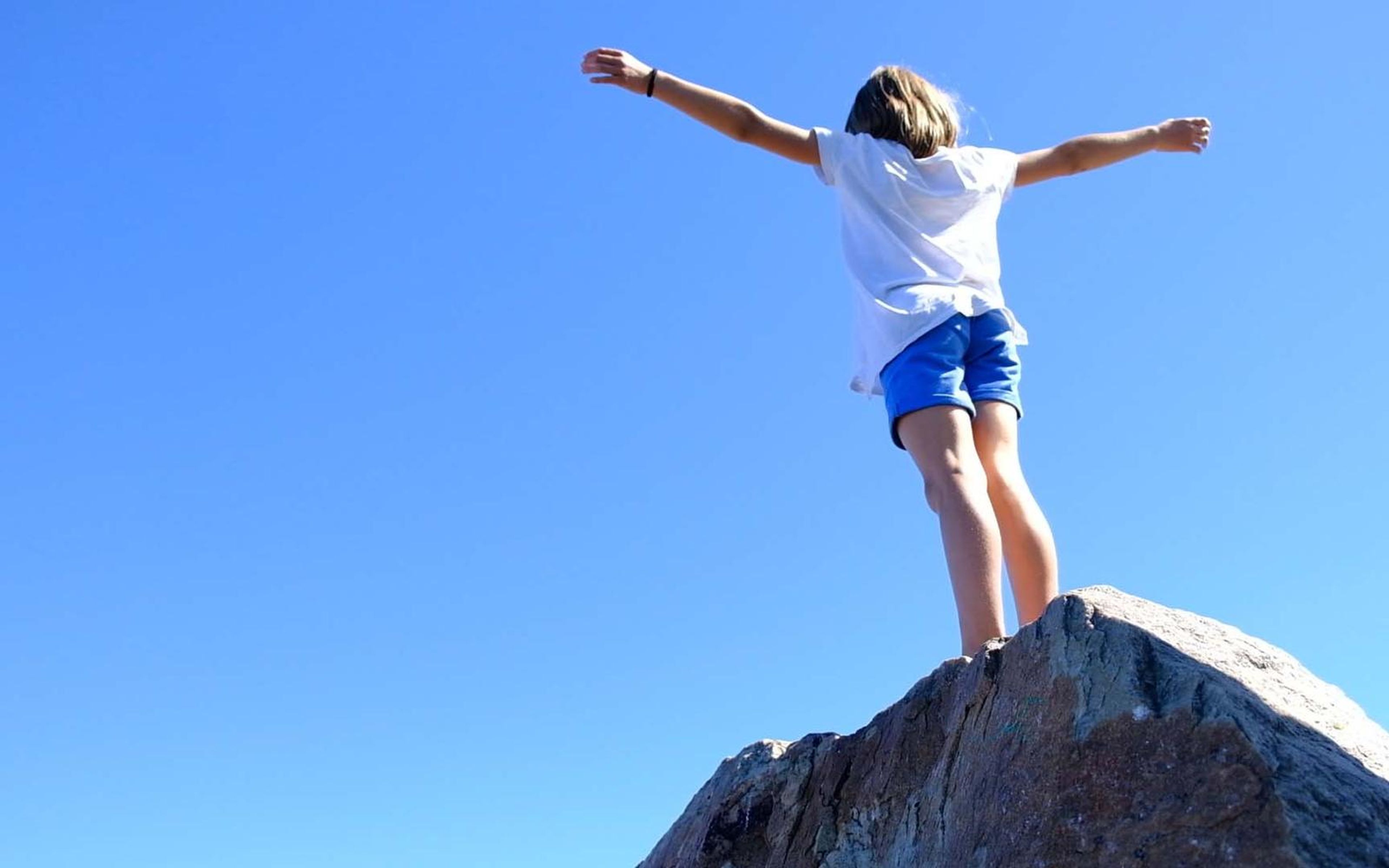 Girl in a white top and blue shorts standing on a rock with arms outstretched against a deep blue sky