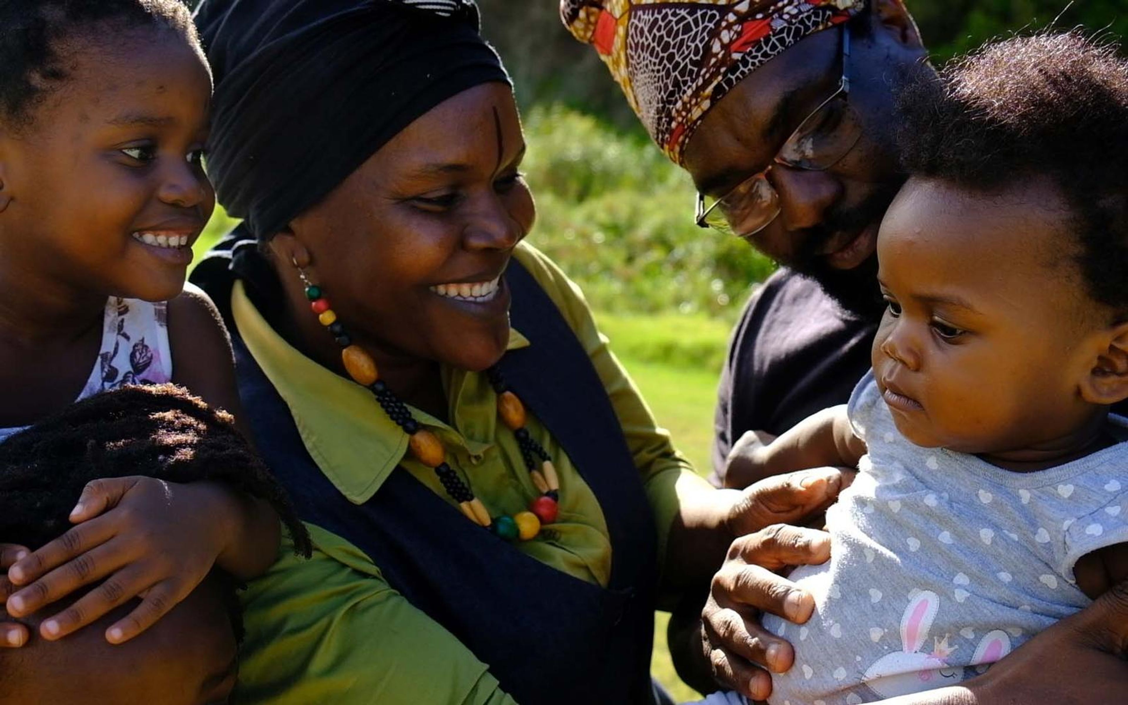 Smiling group outdoors in bright light: a woman with a beaded necklace, a man in glasses, a girl and a baby