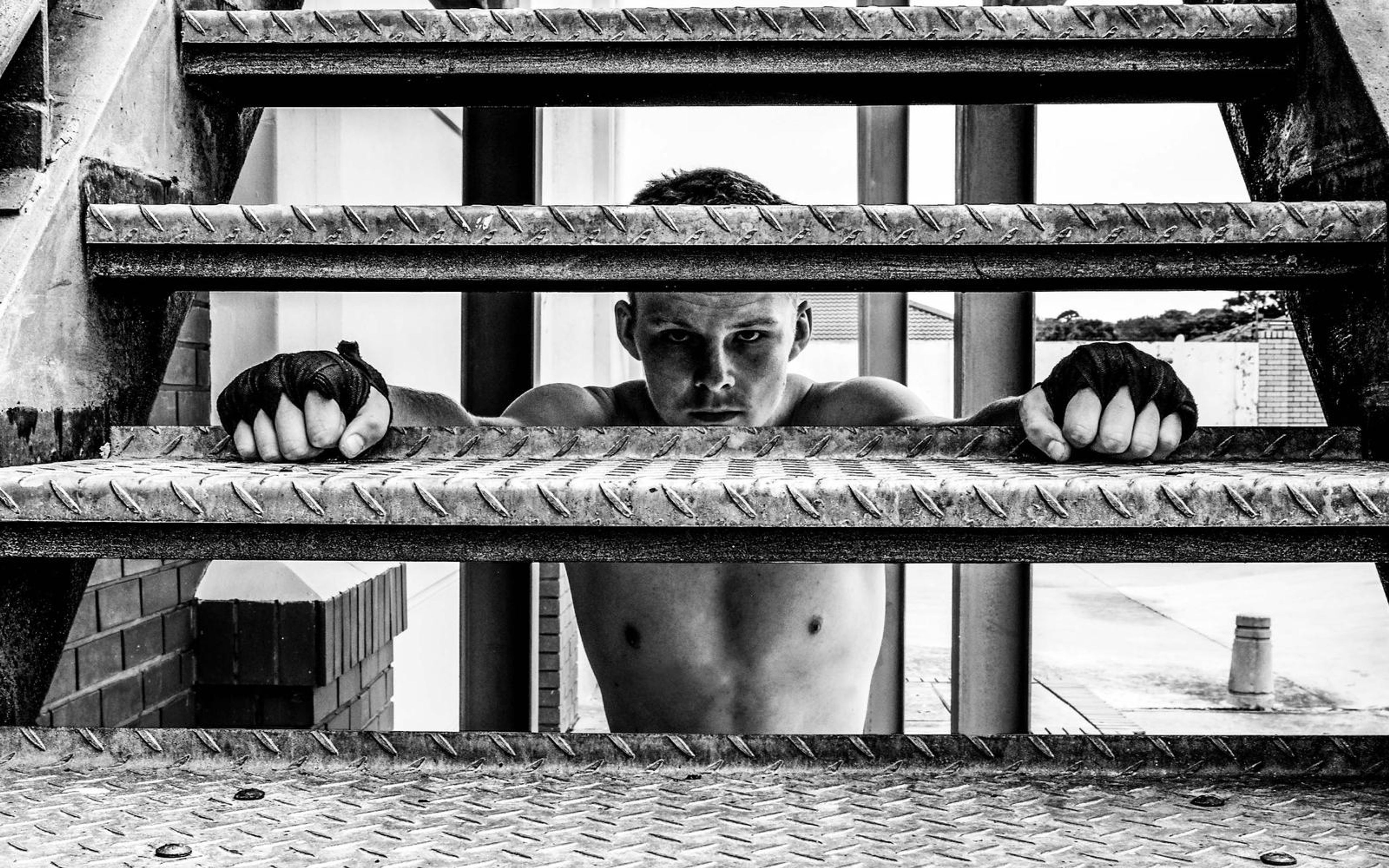 Black and white view of a man's face and wrapped hands gripping a steel stair step, looking through the gap