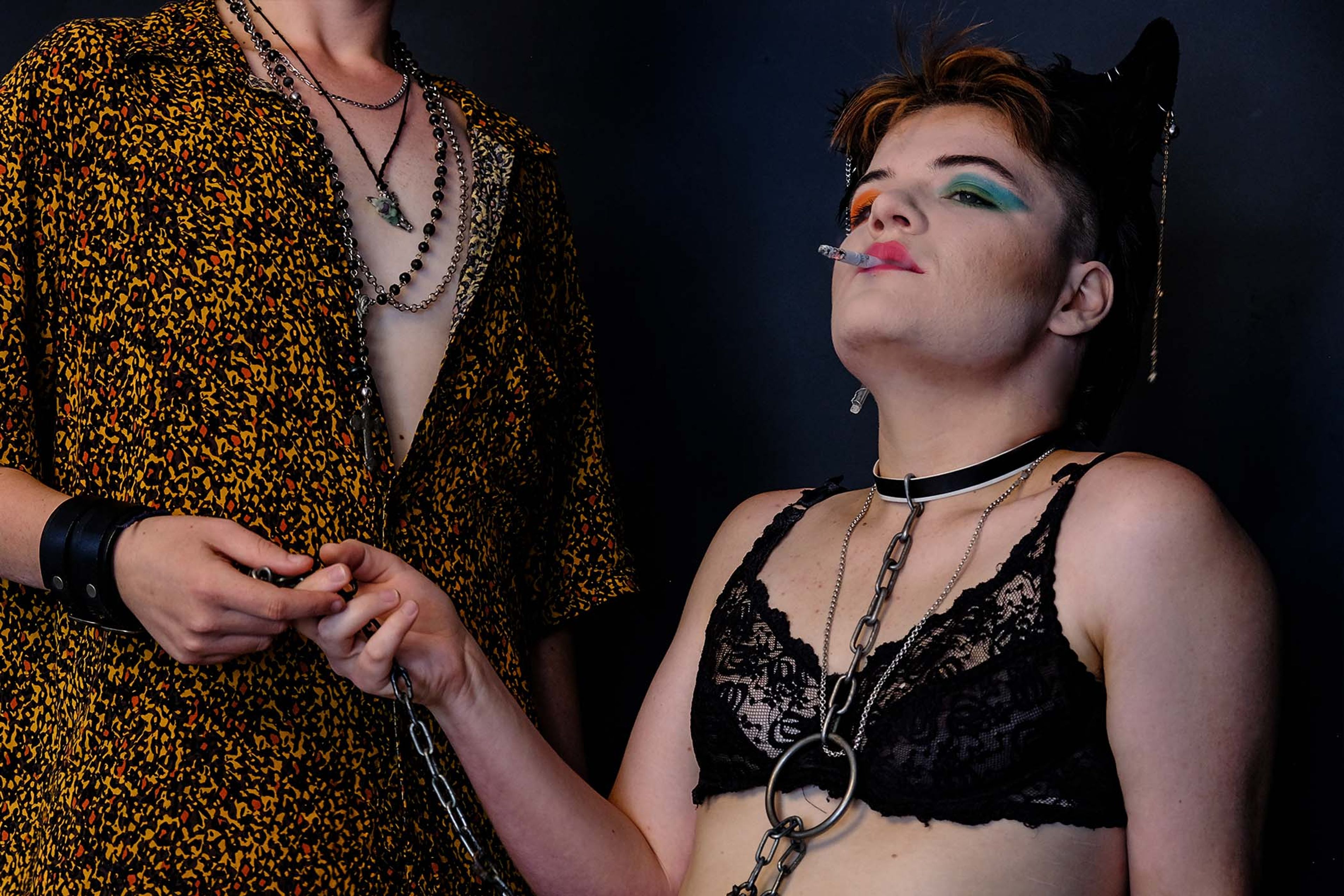 Portrait of a couple, one seated in a lace bra and chain choker holding a cigarette, the other standing beside in a patterned shirt