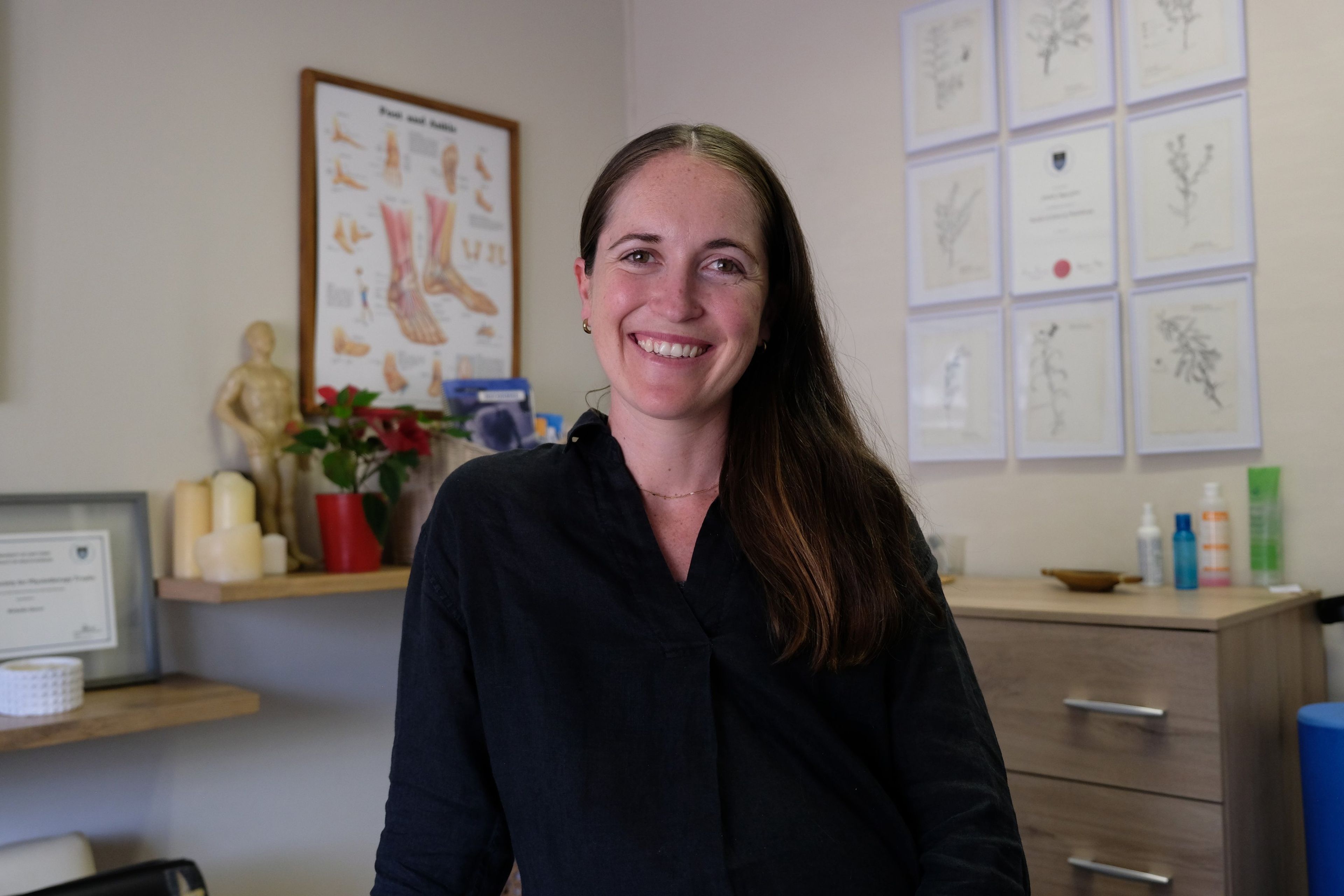 Portrait of a team member in a black shirt smiling in a treatment room with anatomy charts and certificates on the wall
