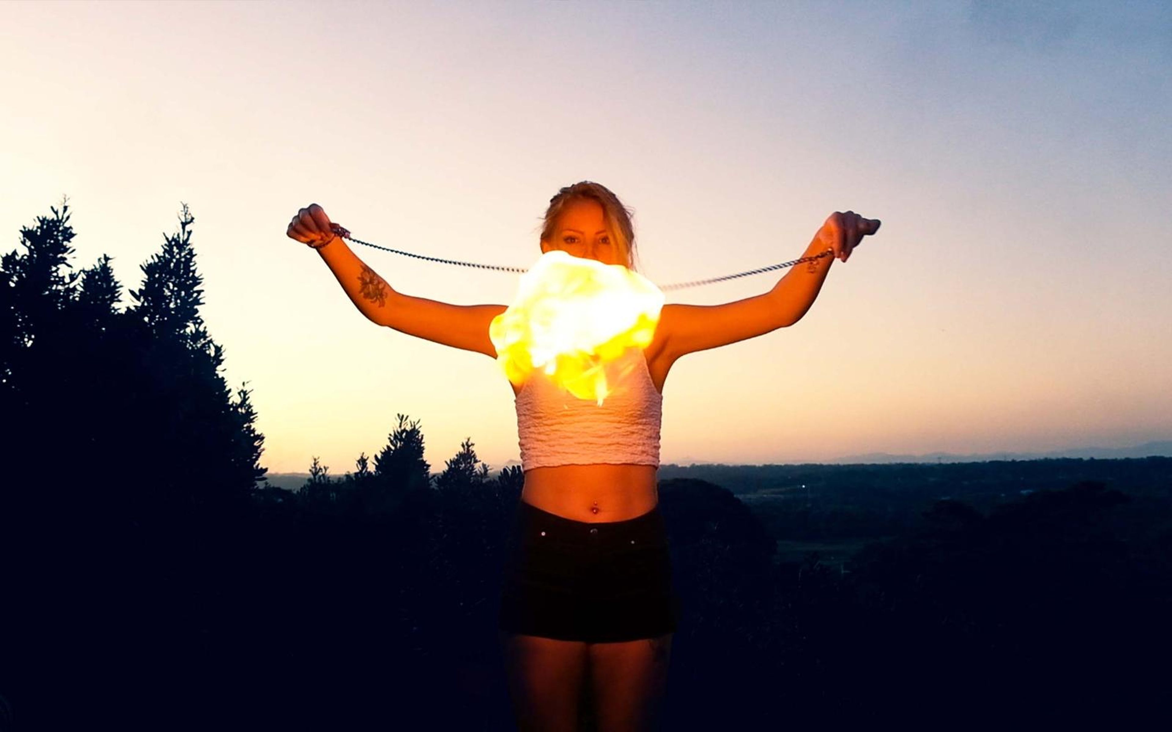 Performer holding a flaming chain across her chest with arms spread, a treeline against a fading sky