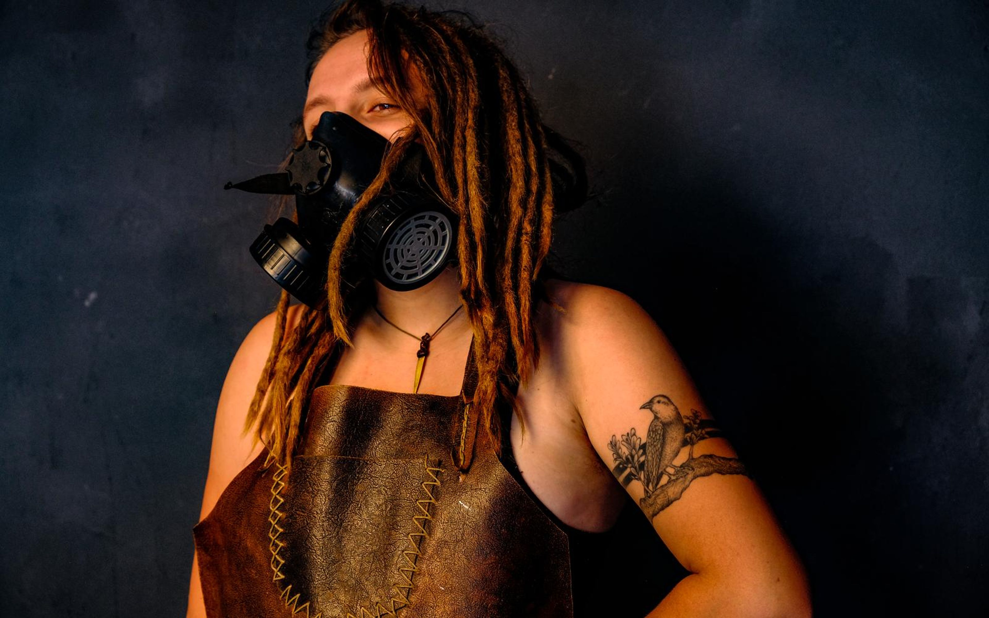 Woman with dreadlocks in a spiked respirator mask and leather apron, lit in warm orange against a dark wall