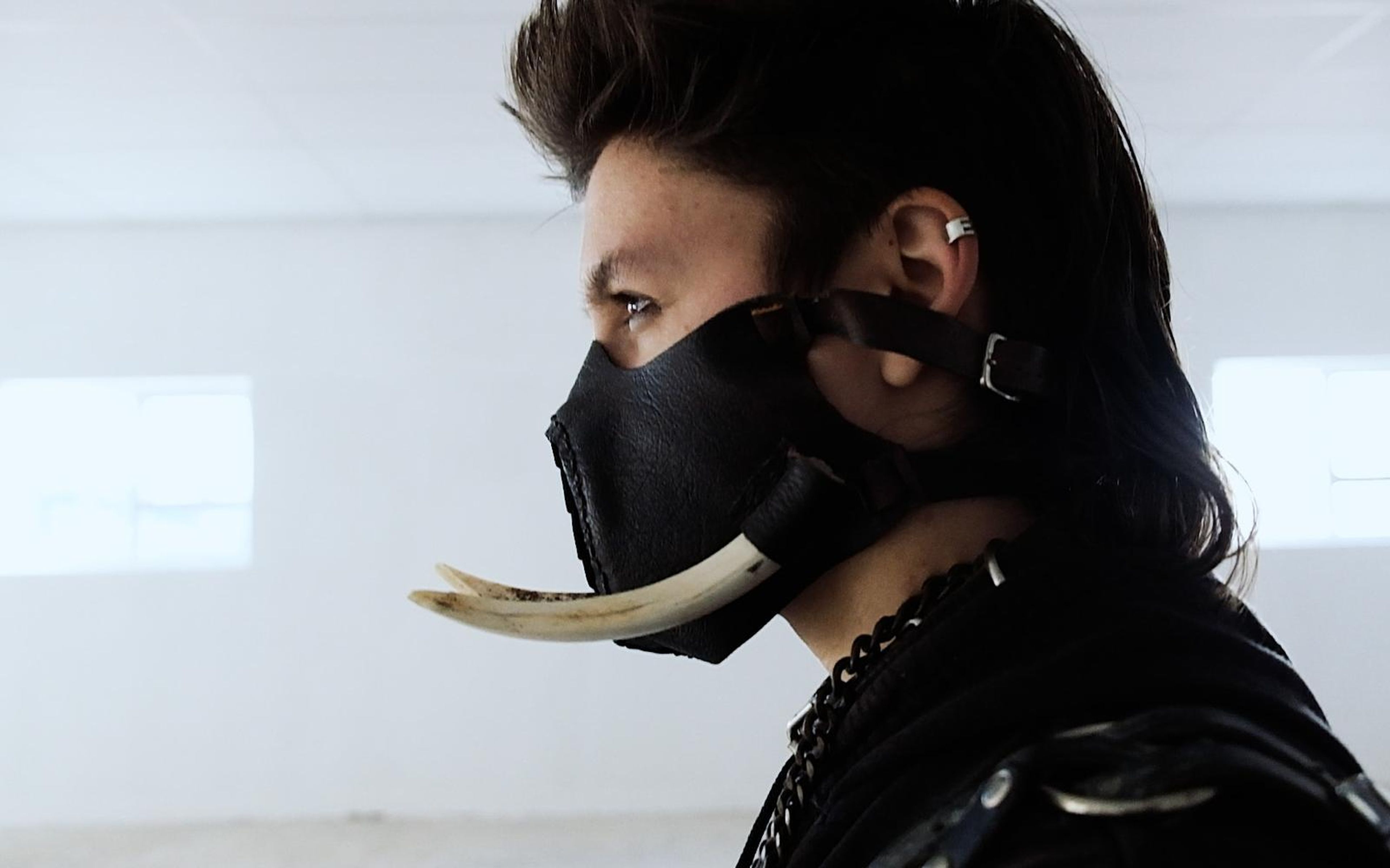 Side profile of a masked figure in a black leather mask with a long curved tusk, white wall and windows behind