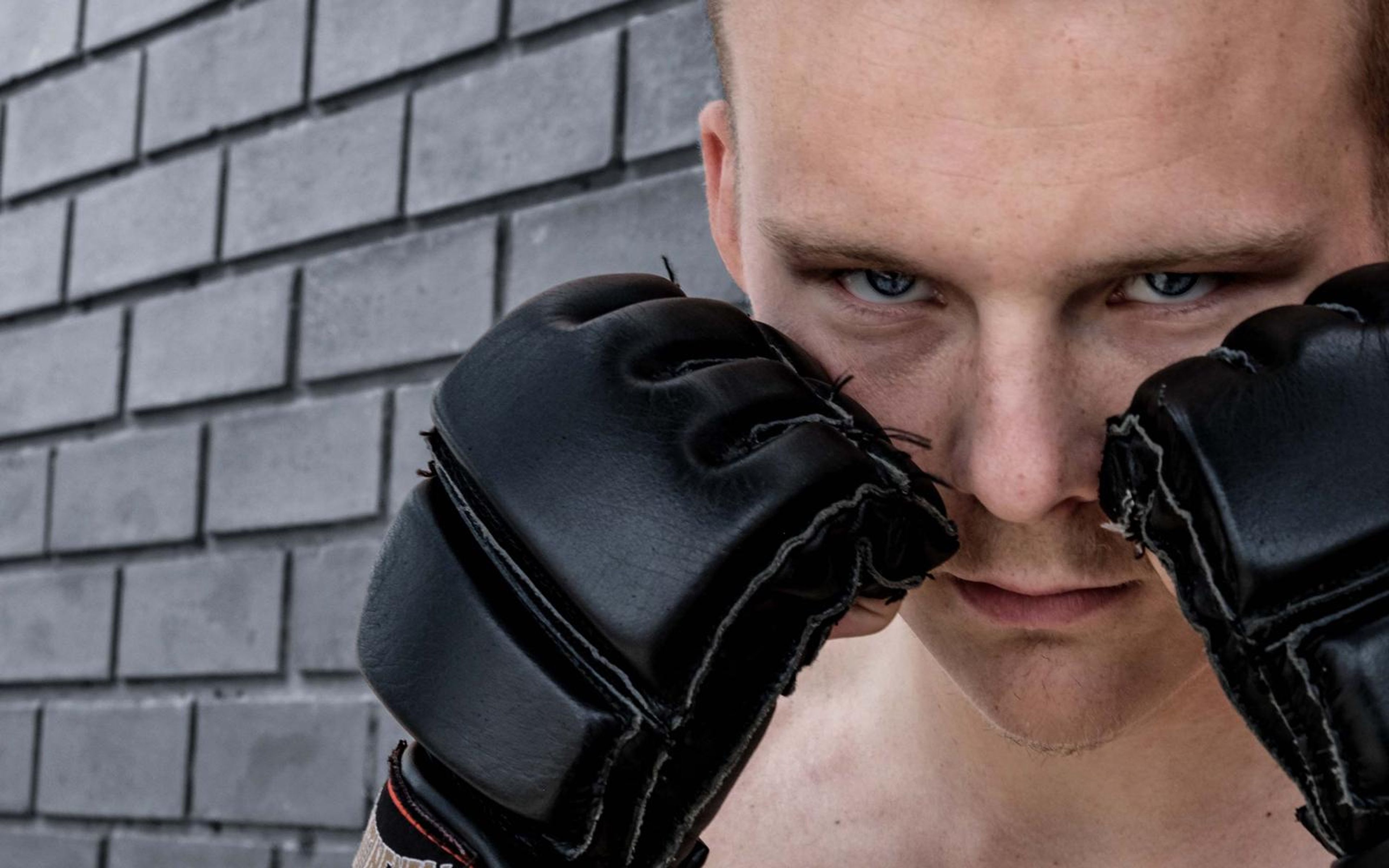 Fighter peering over two MMA gloves held up to his face against a grey brick wall