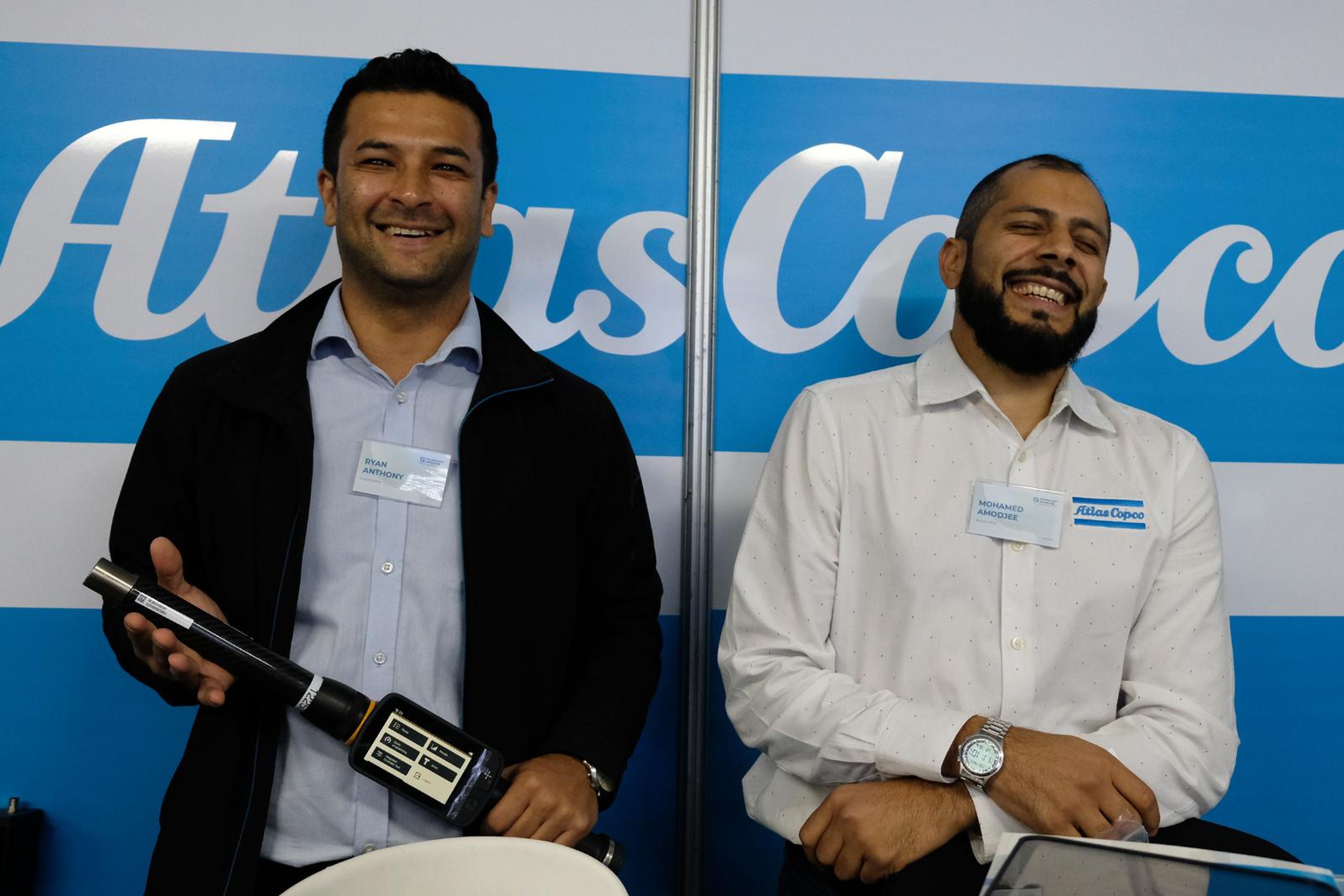 Two Atlas Copco team members smiling beside a handheld tool at their stand