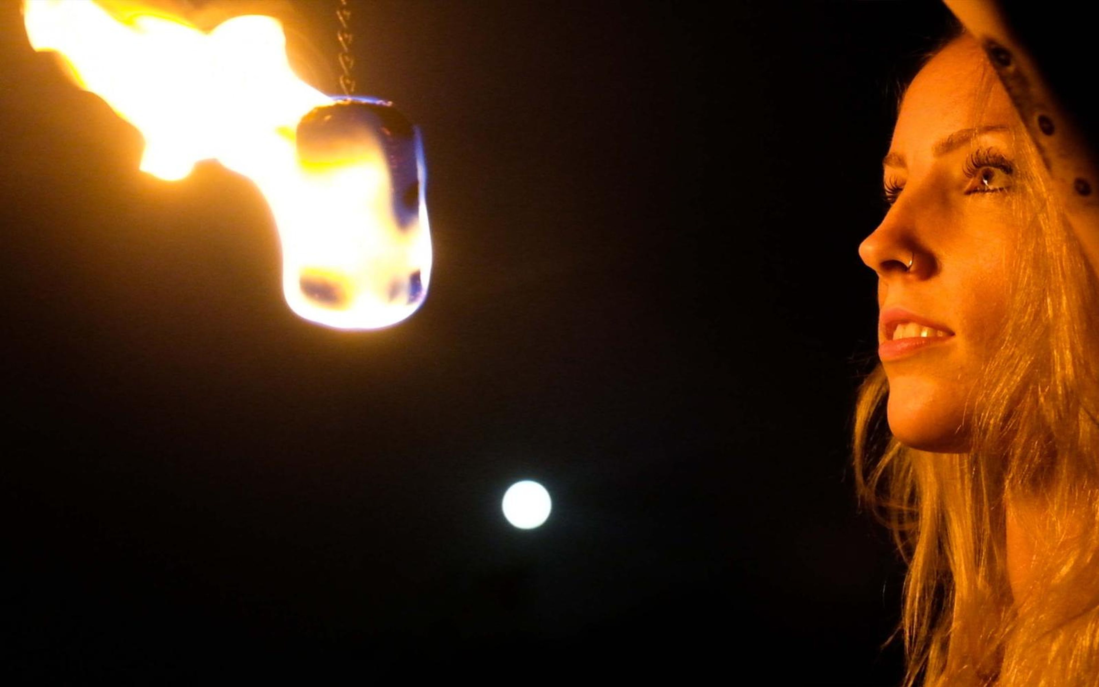 Close-up of a woman's face in warm firelight beside a large flame, with a bright spot of light below
