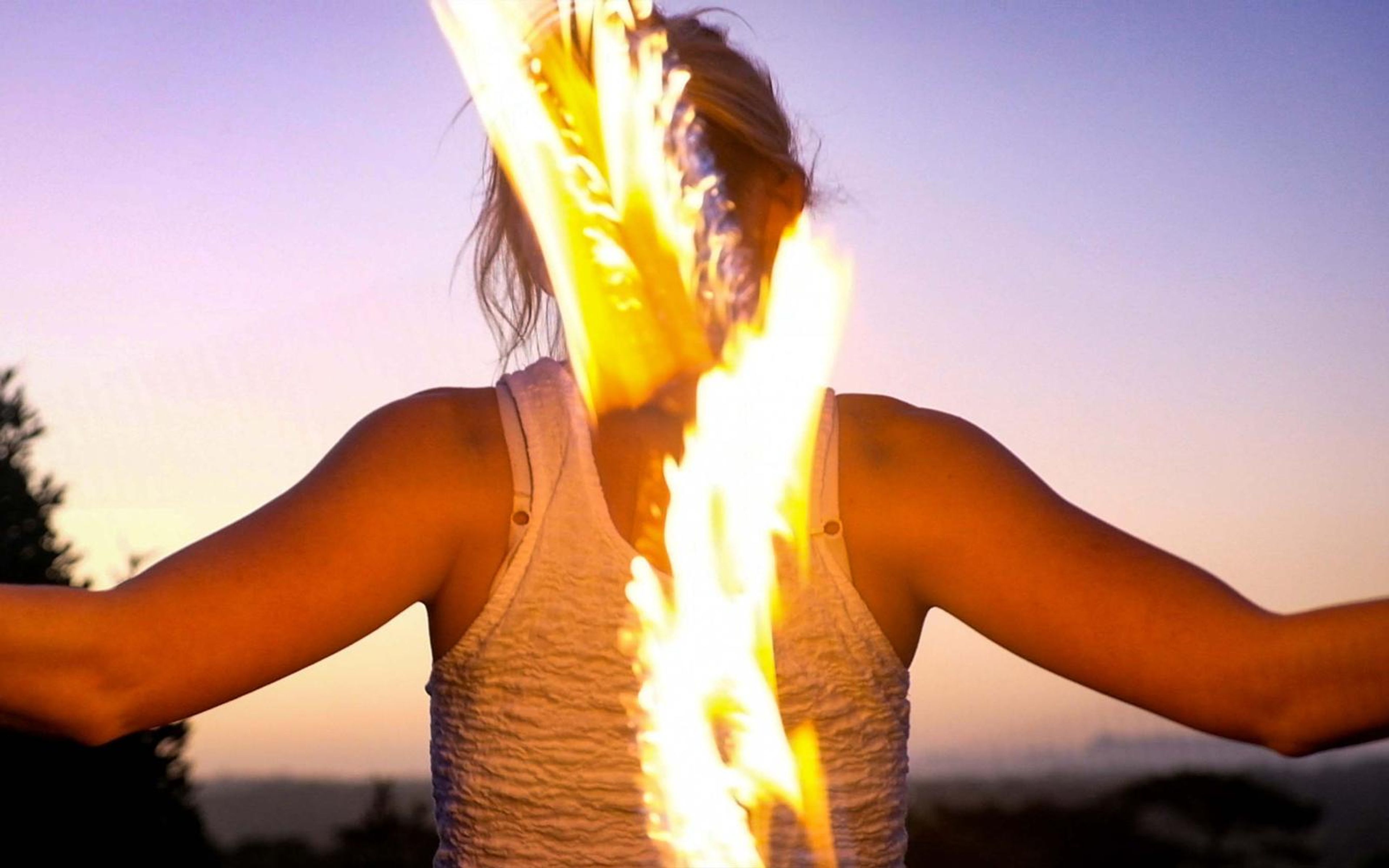 Woman from behind with arms out and tall flames rising either side of her head against a violet sky