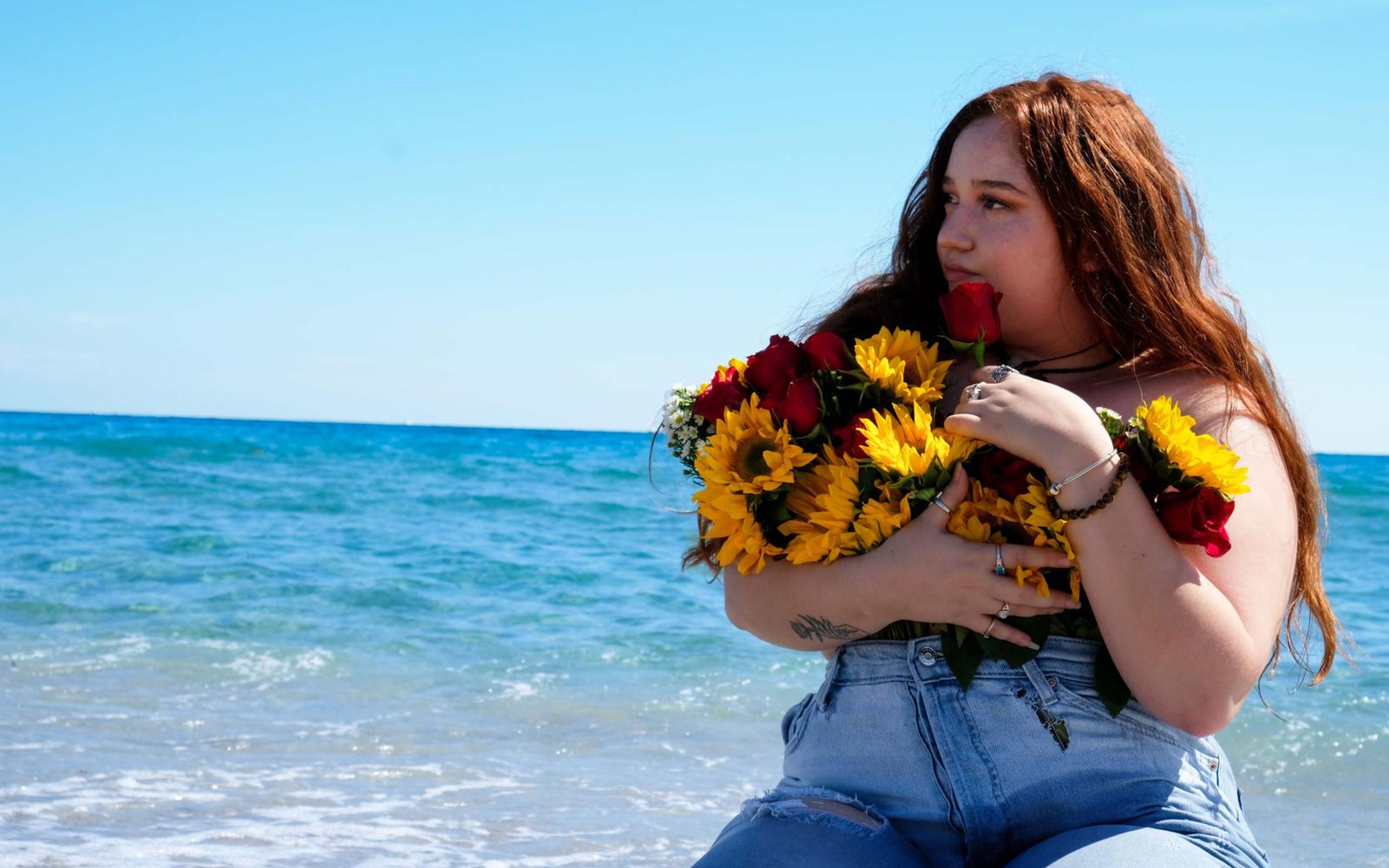 Woman seated in shallow surf holding a rose to her lips, hugging a sunflower and rose bouquet, looking out to sea, denim overalls, blue sky