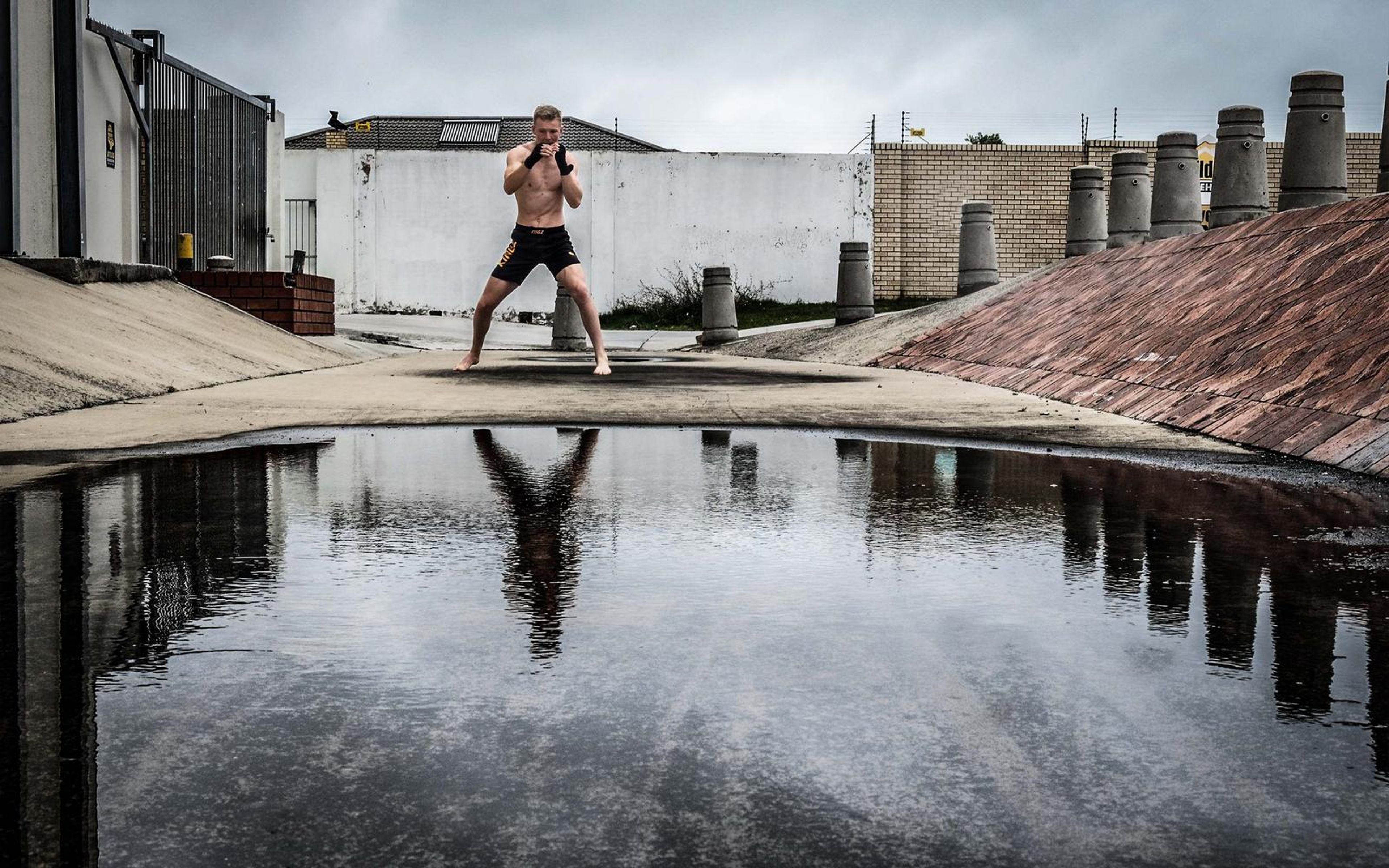 Fighter shadow-boxing beside a flooded puddle with his reflection in the water and chimney stacks beyond
