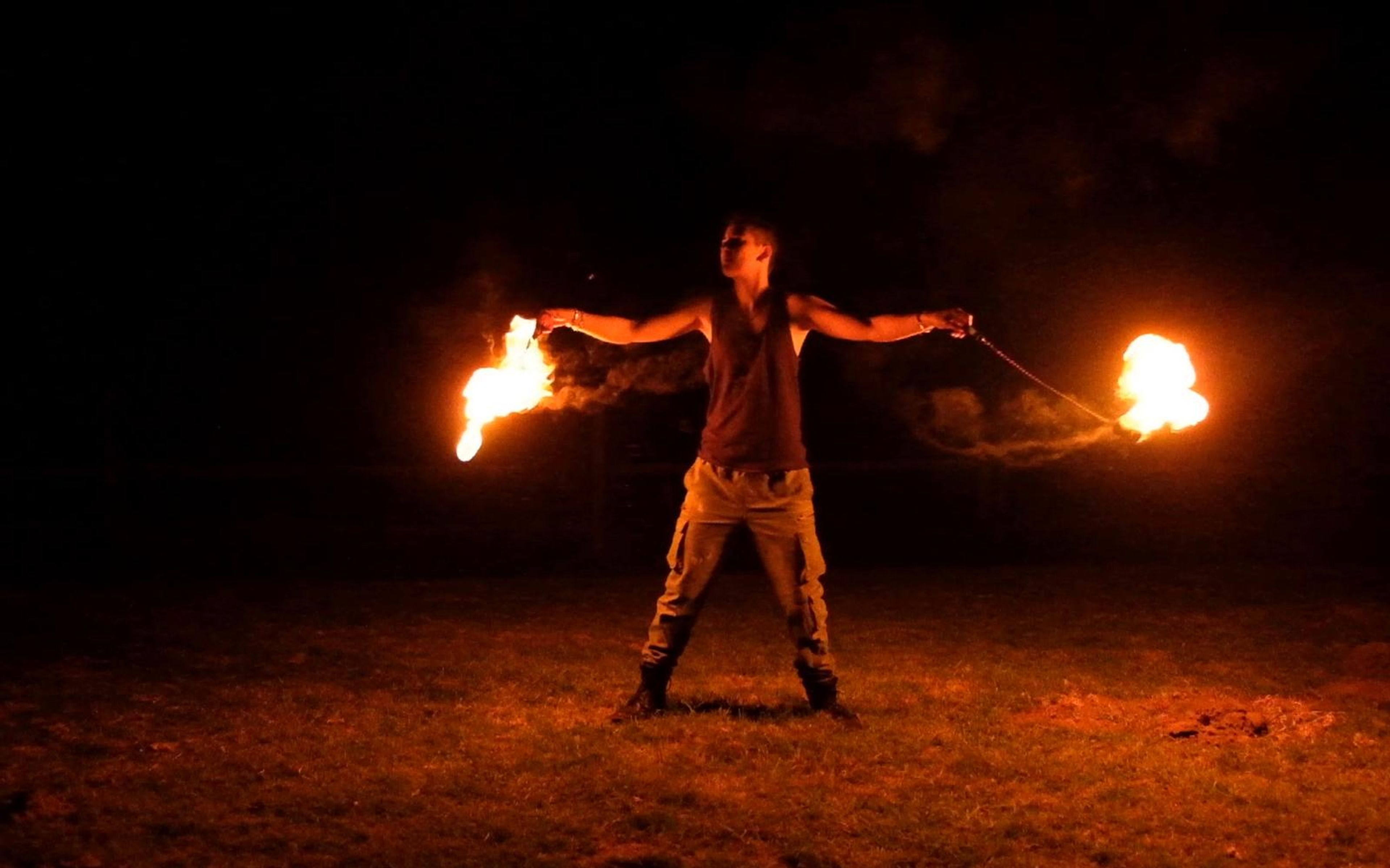 Performer standing with arms wide holding two flaming poi on gravel
