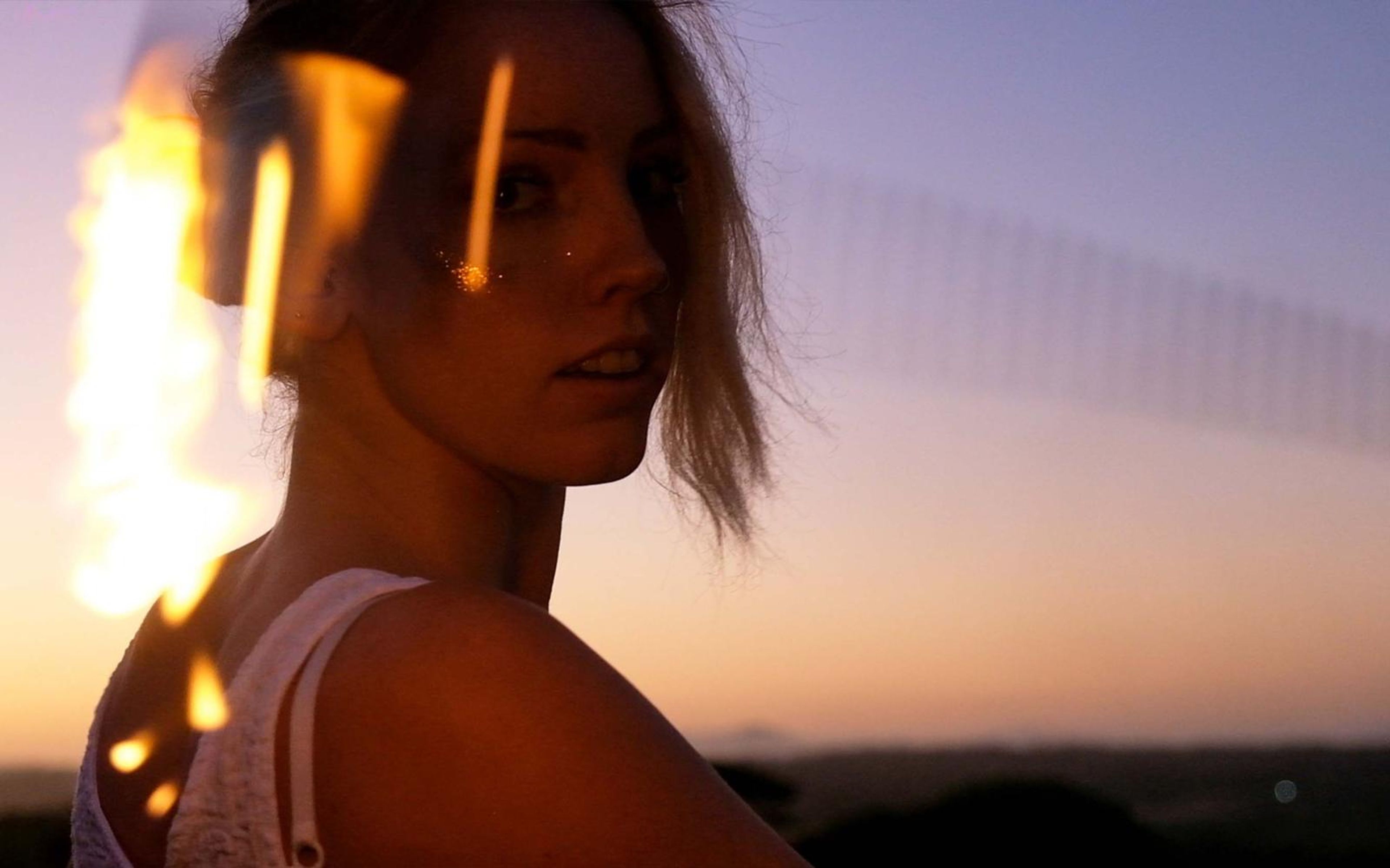 Woman in a white top looking back over her shoulder, flame streaking at left, pink and violet dusk sky