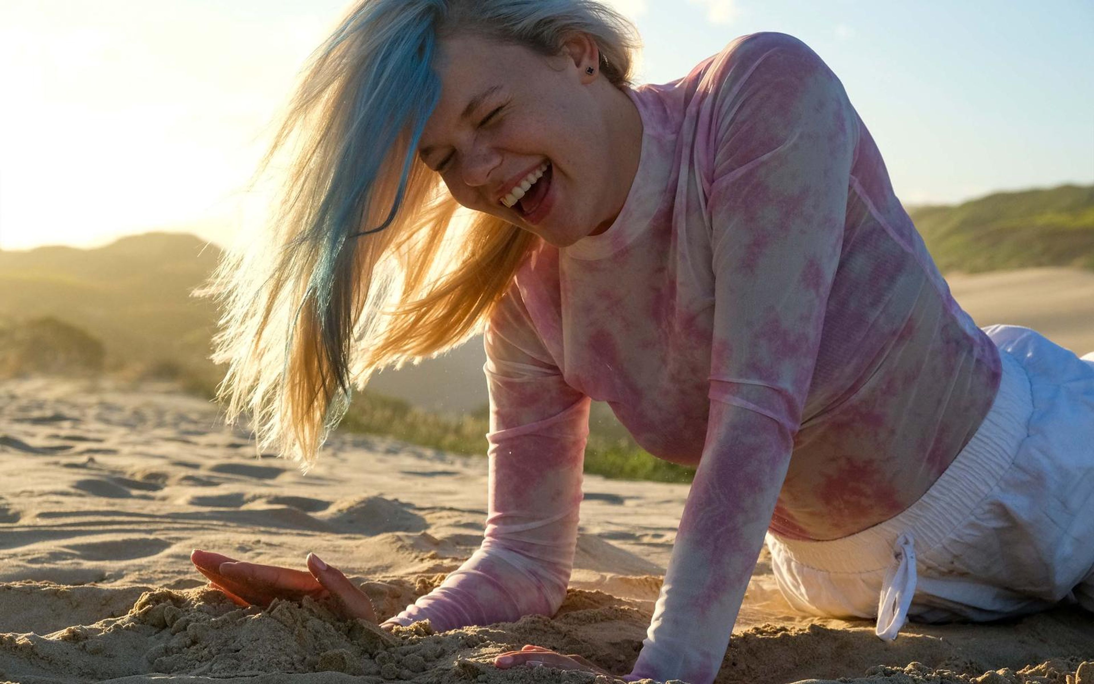 Woman laughing on all fours in the sand with hair blowing across her face, sun behind