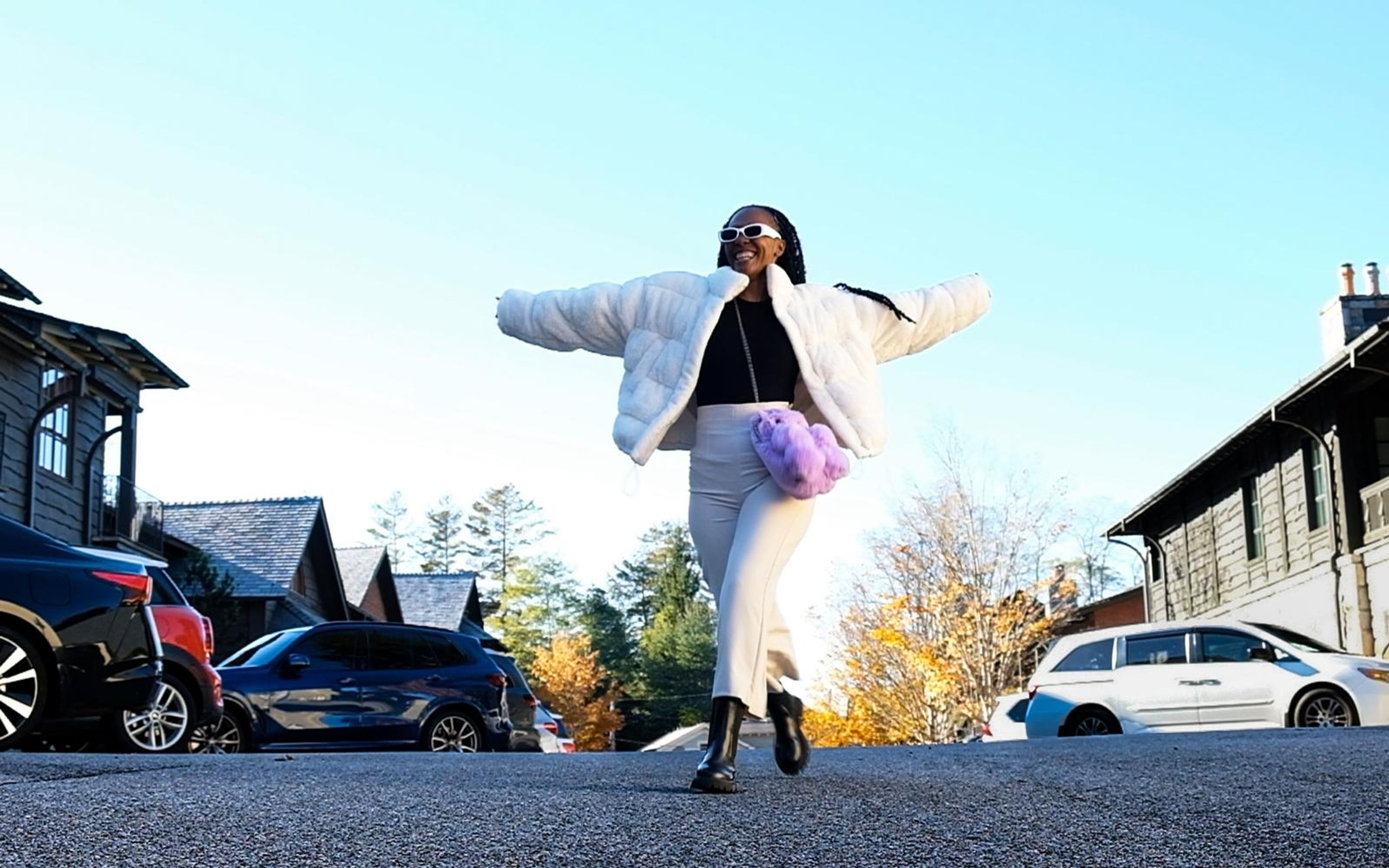Model dancing in the middle of a street, arms wide, white fur jacket, cream trousers, purple fluffy bag, autumn trees