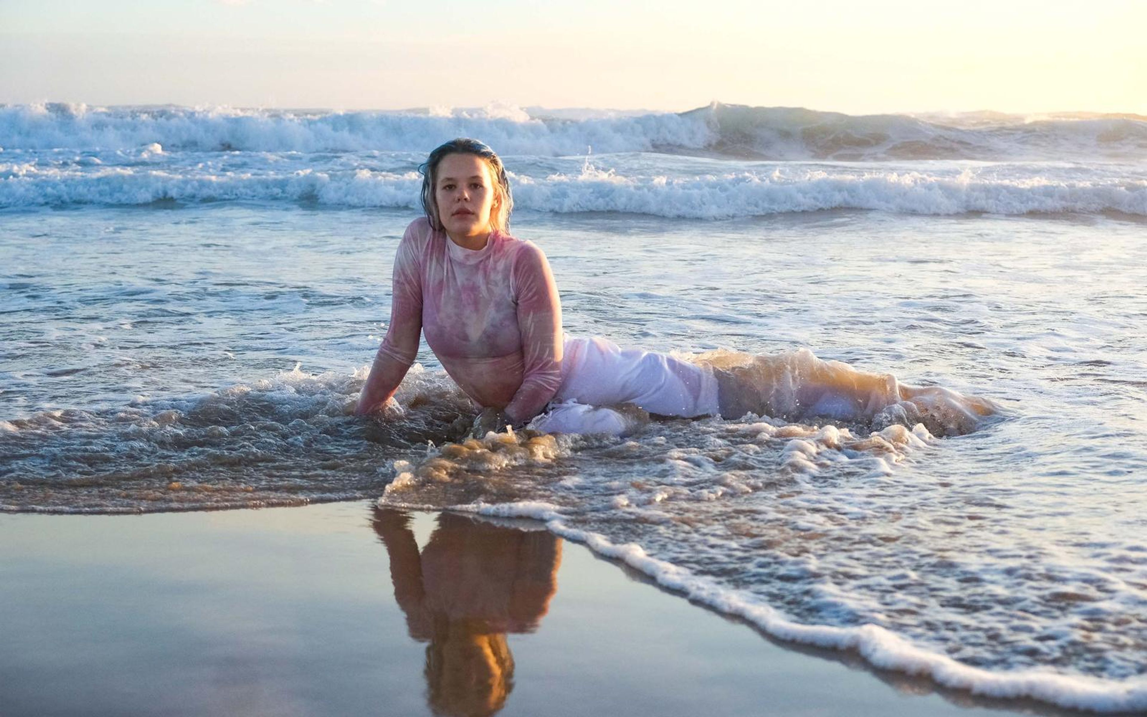 Woman lying in a wide wash of surf with her reflection on glossy sand, pink top, white trousers