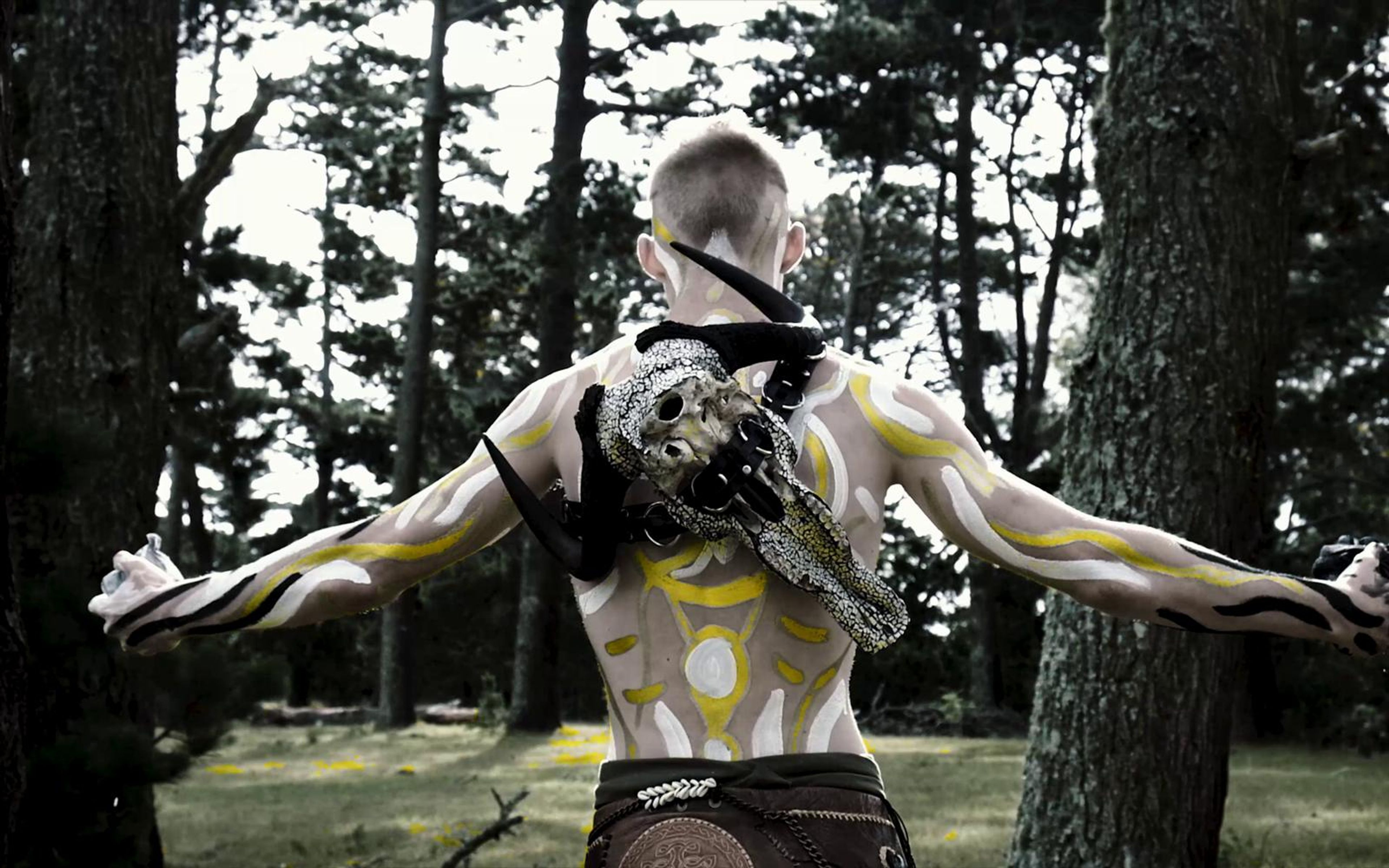 Performer seen from behind with arms stretched wide and a skull on his back in a sunlit pine clearing