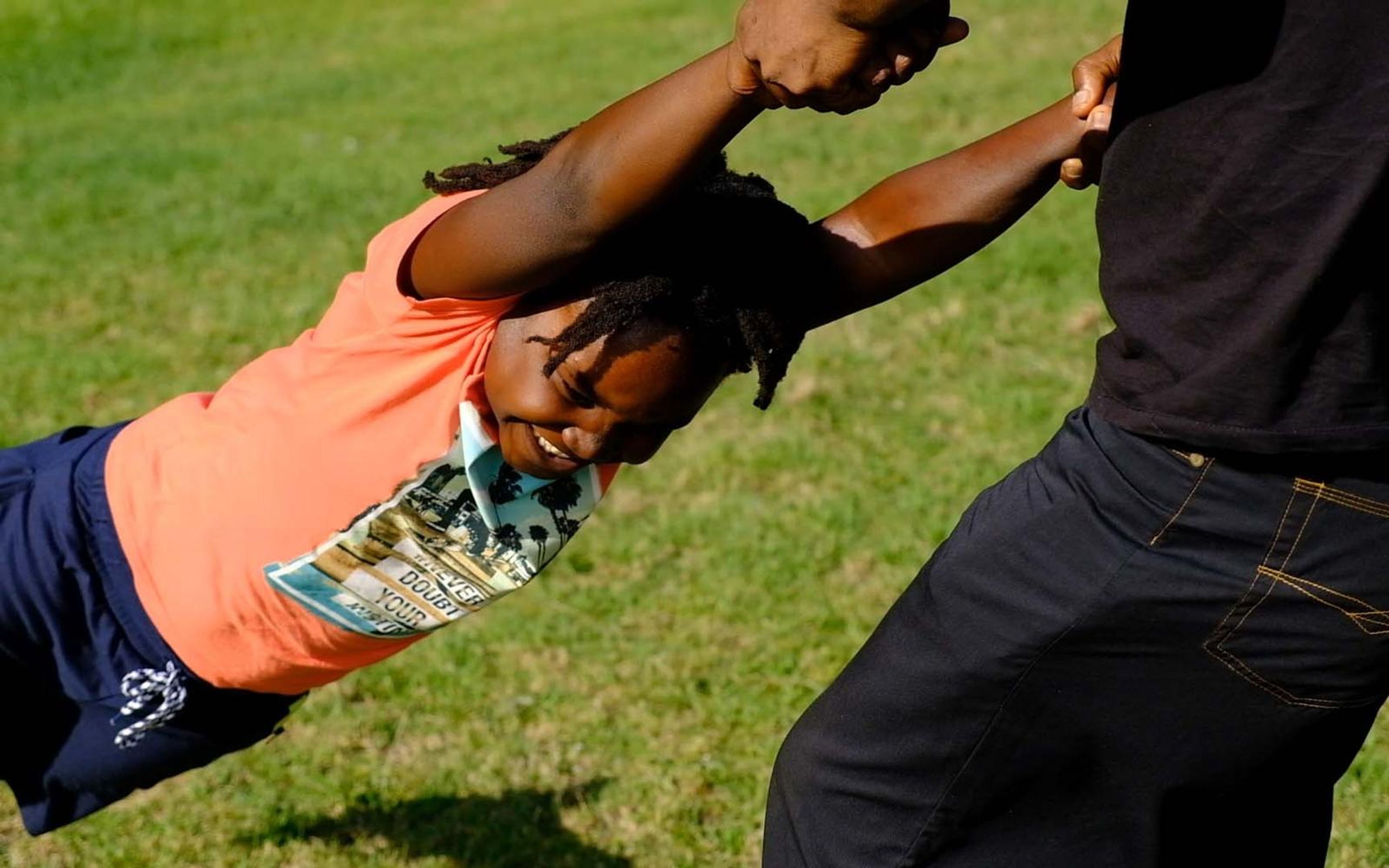 Child in an orange top swung by the hands through the air above green lawn