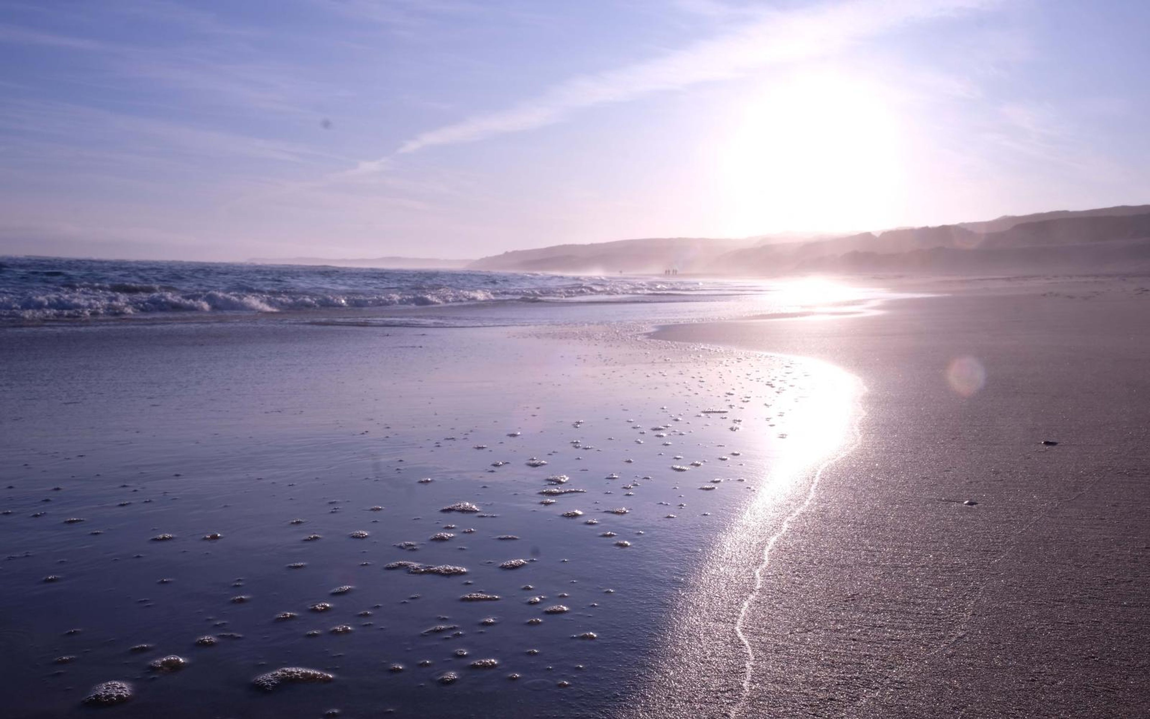 Wet sand mirroring low sun in a bright reflection line along the shore, hills in haze behind