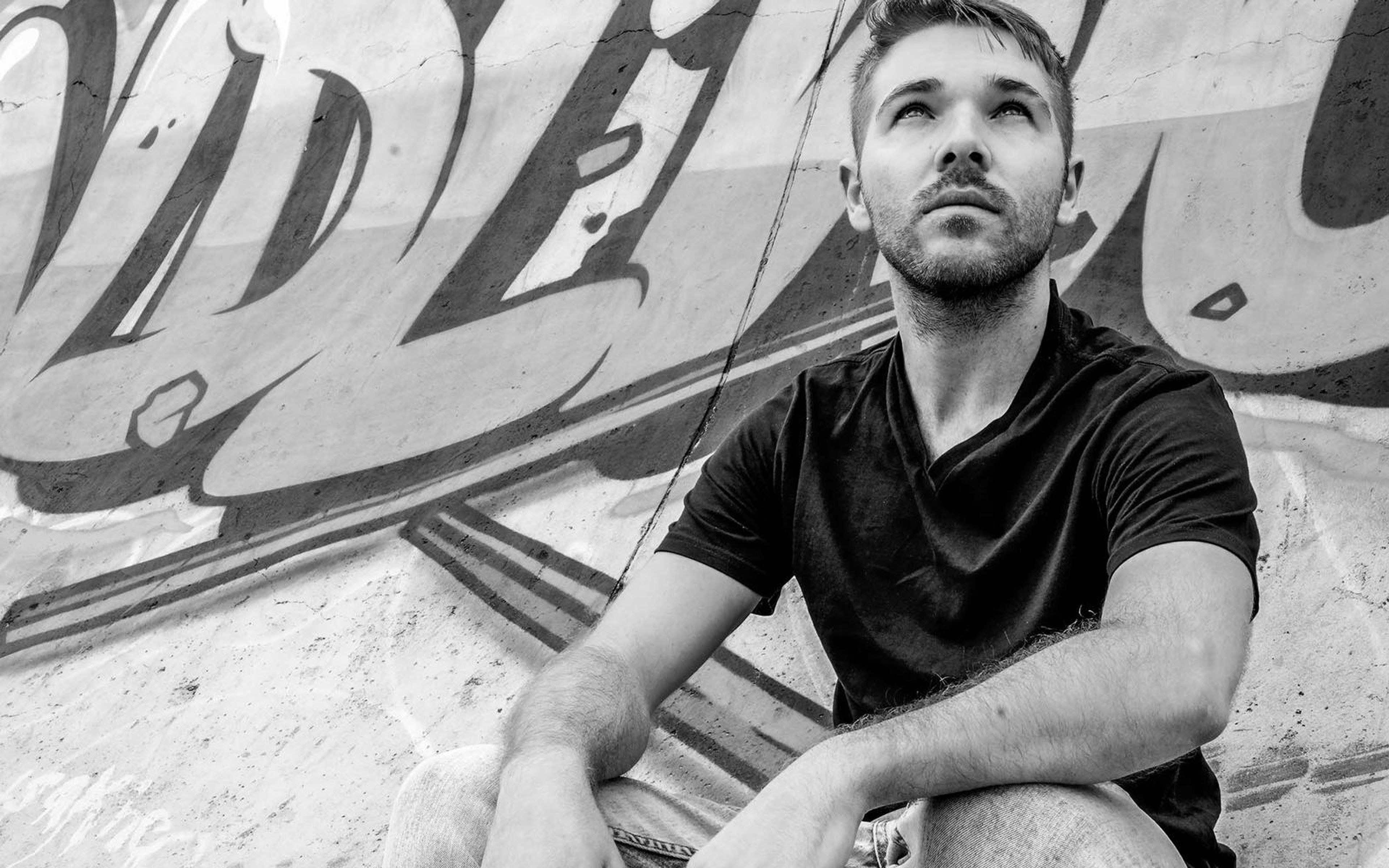 Black and white portrait of a man in a dark tee looking upward in front of graffiti lettering