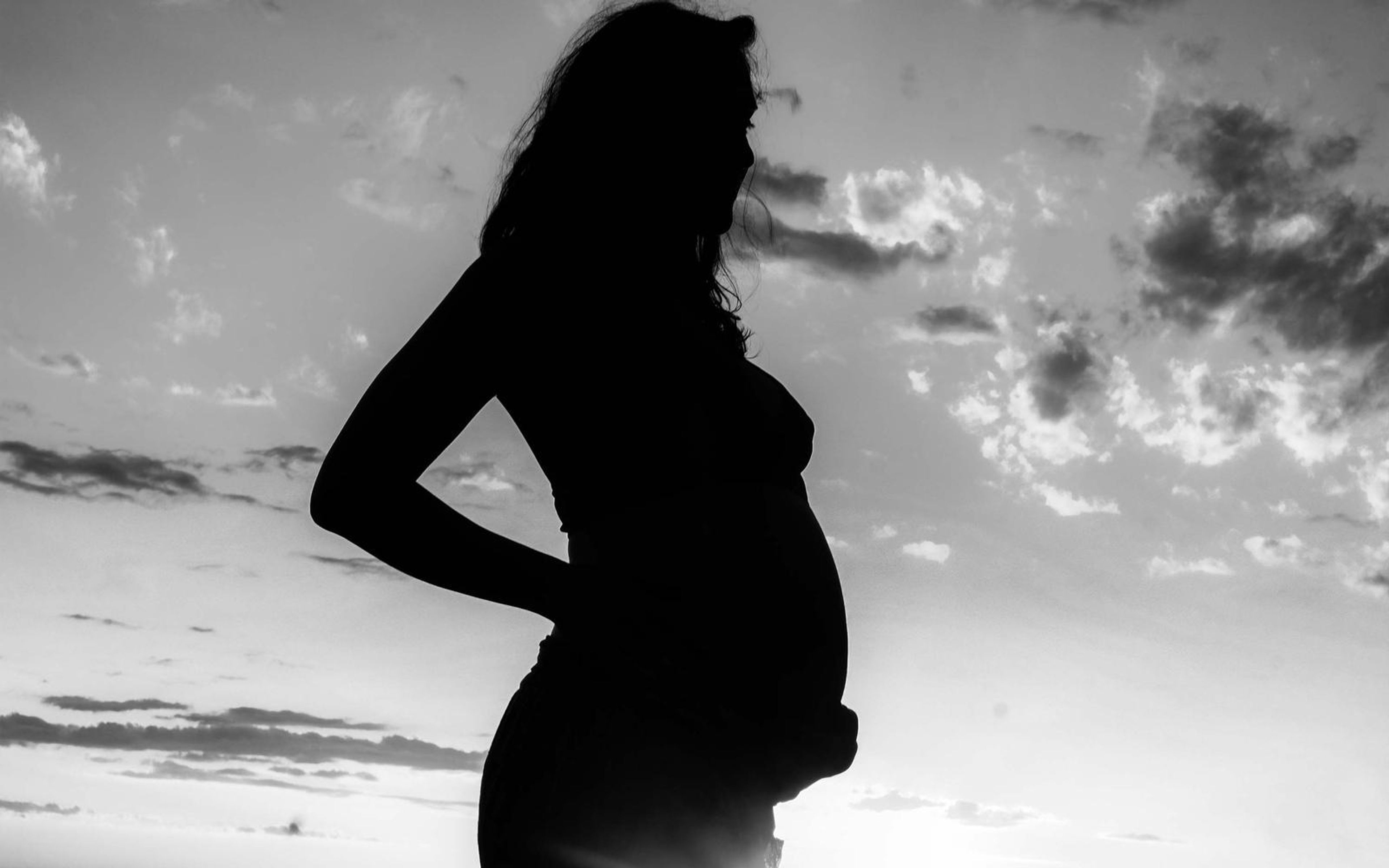 Black and white silhouette of a pregnant woman, hand on her back, bright sky with dark clouds